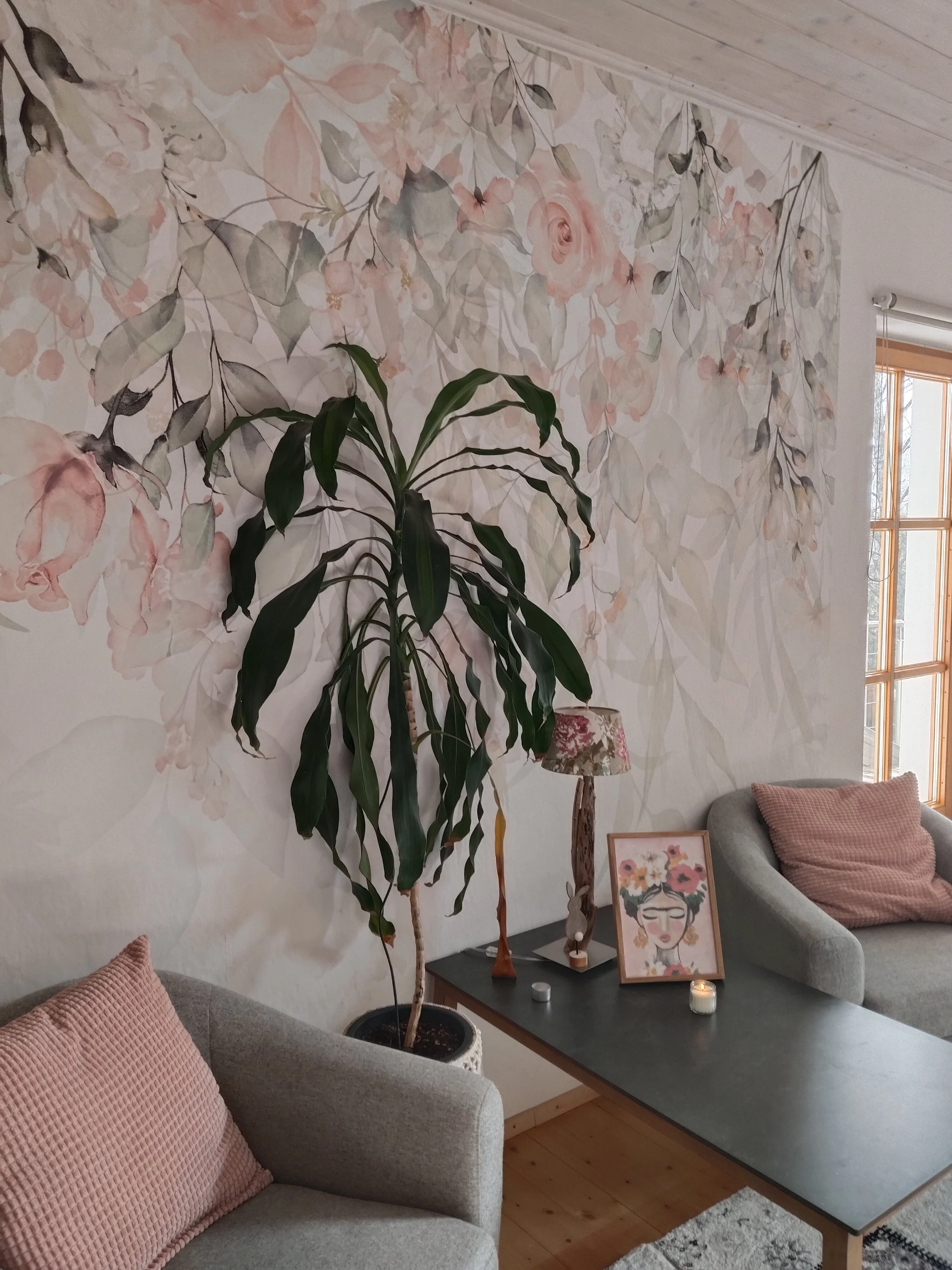 Gemälde einer Frau mit Brille und Blumen im Haar auf einem Tisch, neben einer großen grünen Zimmerpflanze in einem Wohnzimmer mit floralem Wandbild, einem Bild im Rahmen, einer Tischlampe, Kerzen und einem Sofa