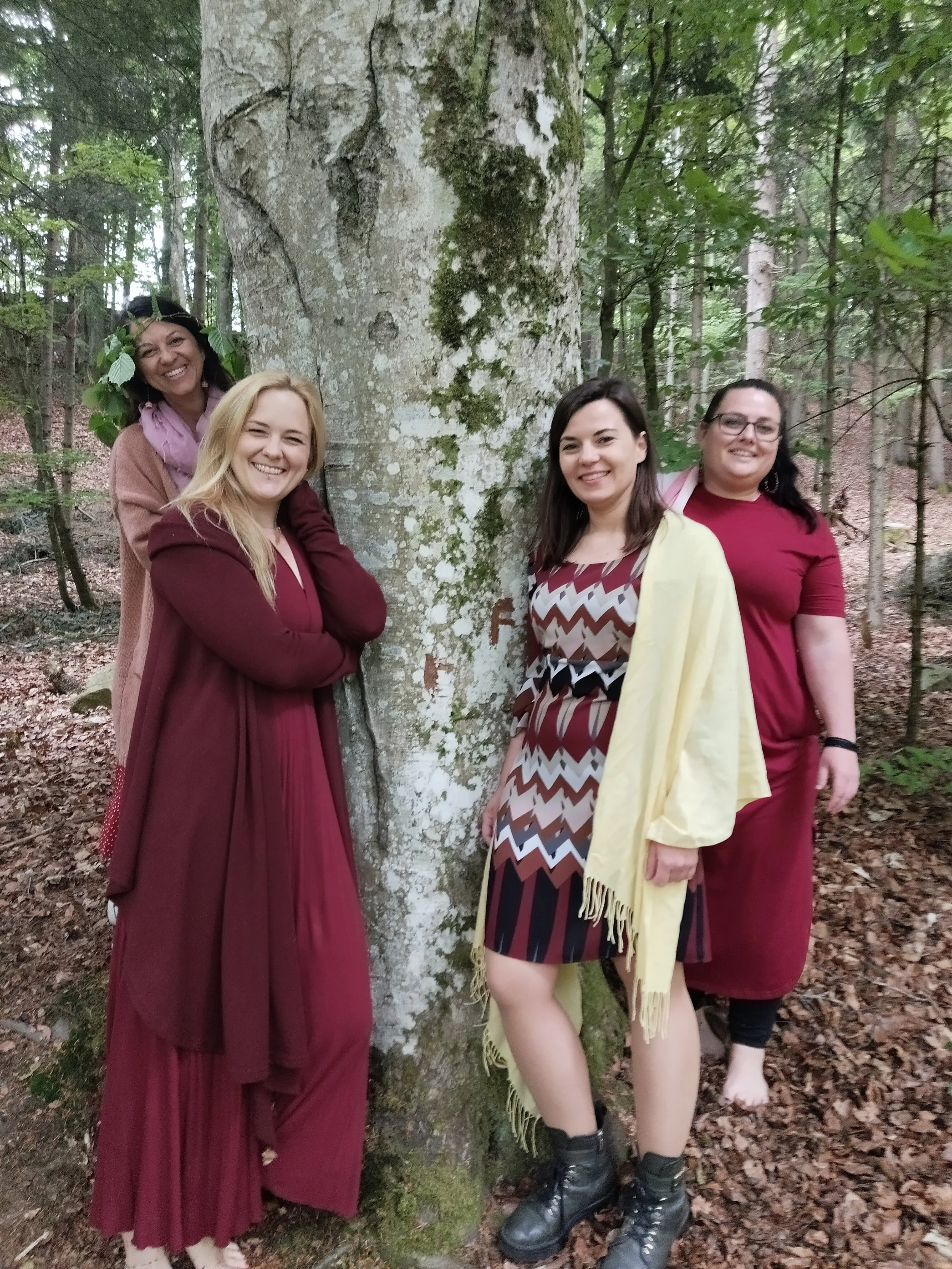 Vier Frauen lachen, die um einen Baum im Wald stehen, tragen bunte Kleider und Schuhe, einige tragen Schuhe, eine Frau ist barfuß.