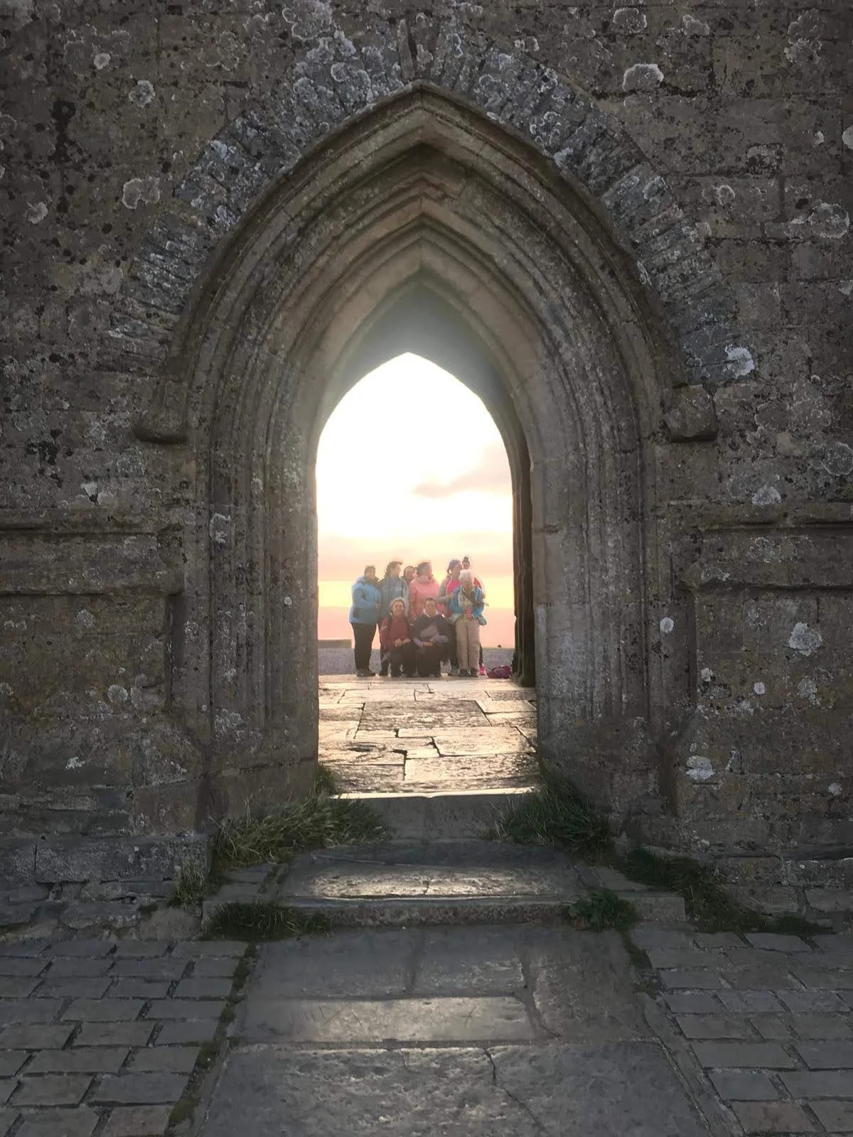 Eine Gruppe von Menschen steht vor einer alten, gotischen Fensteröffnung in einer Steinmauer, bei Sonnenuntergang.