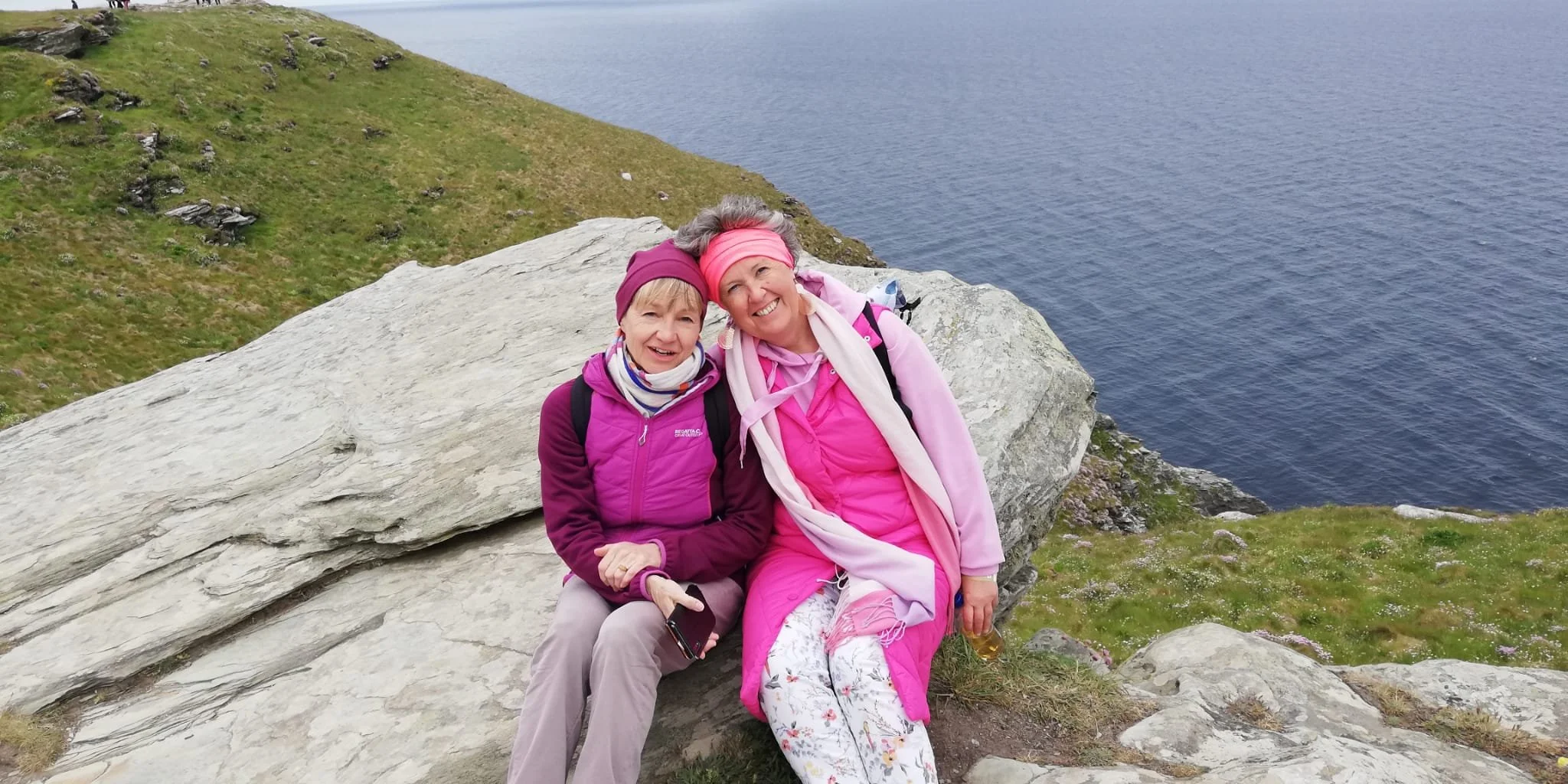 Zwei lachende Frauen sitzen auf einem großen Stein an der Küste, umgeben von grünen Hügeln mit Blick auf das Meer. Sie tragen pinke und lila Kleidung und Kopfbedeckungen.
