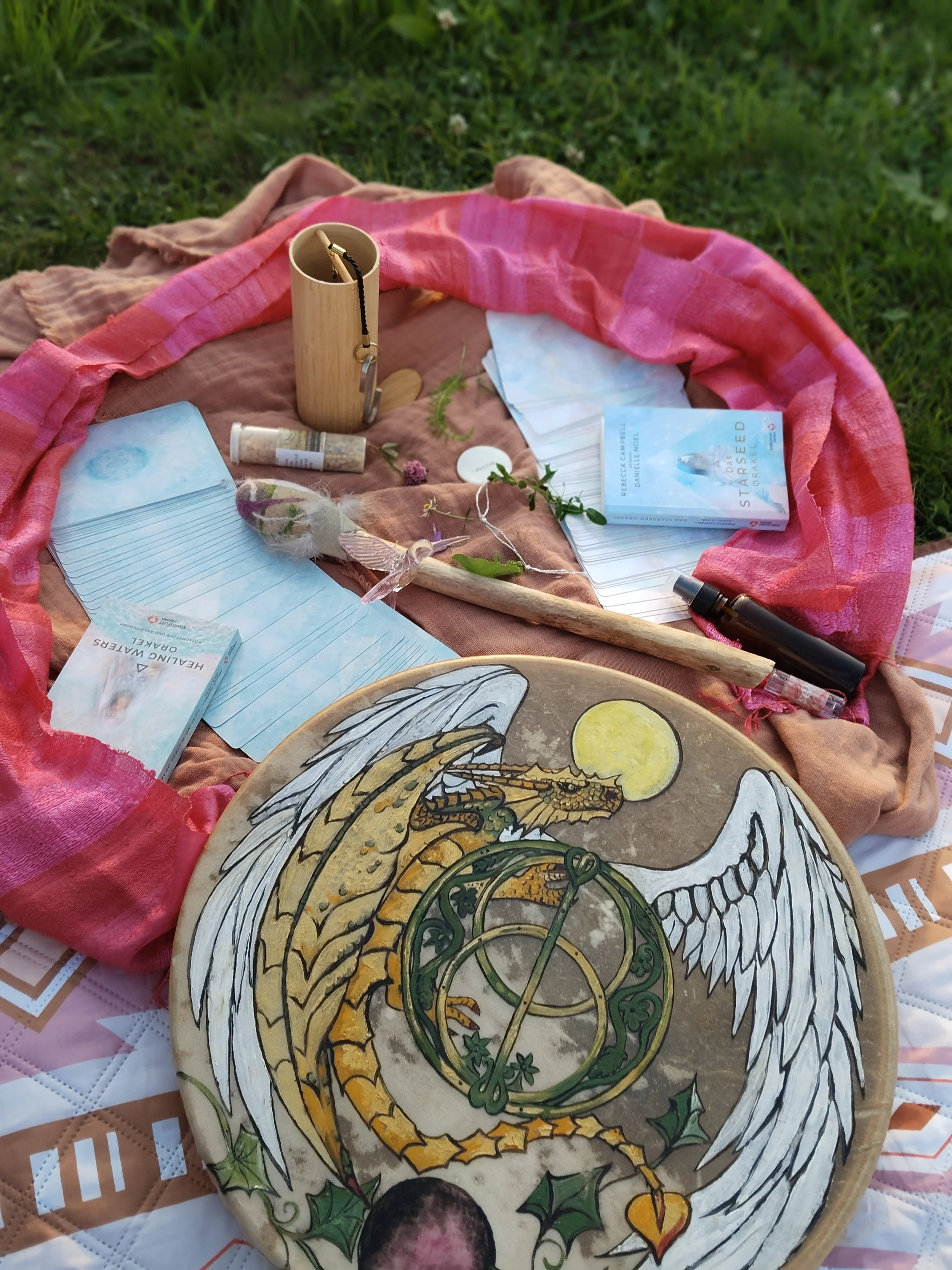 Ein horoskopischer Drachen und ein Kreis auf einem terrakottafarbenen Teller, umgeben von Tarot-Karten, einem Geschenk mit Federn, einer kleinen Flasche, einem Spray und einer Mappe, auf einem roten Tuch im Freien.