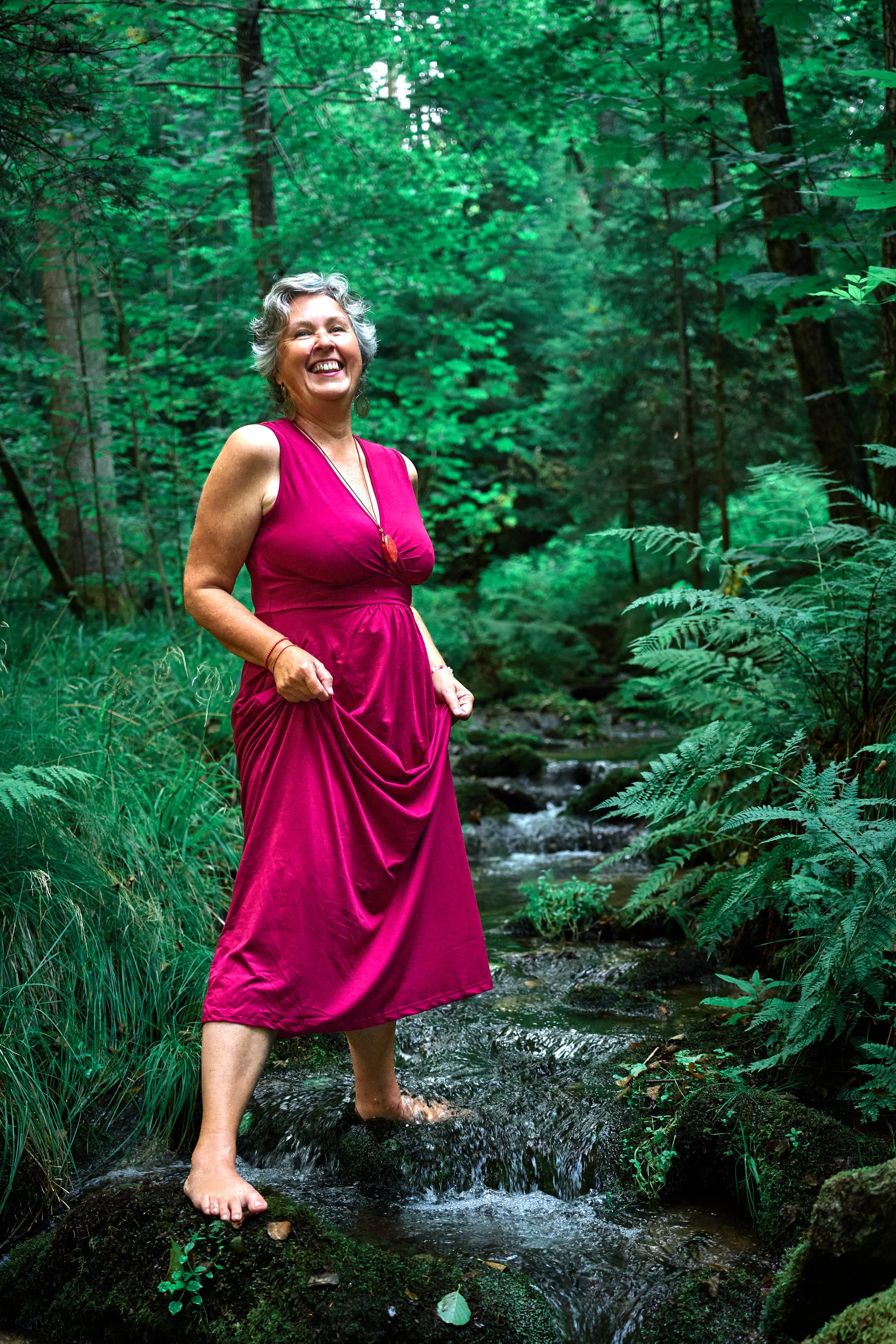 Eine lächelnde ältere Frau in einem pinken Kleid, die barfuß in einem Bach in einem grünen Wald steht.