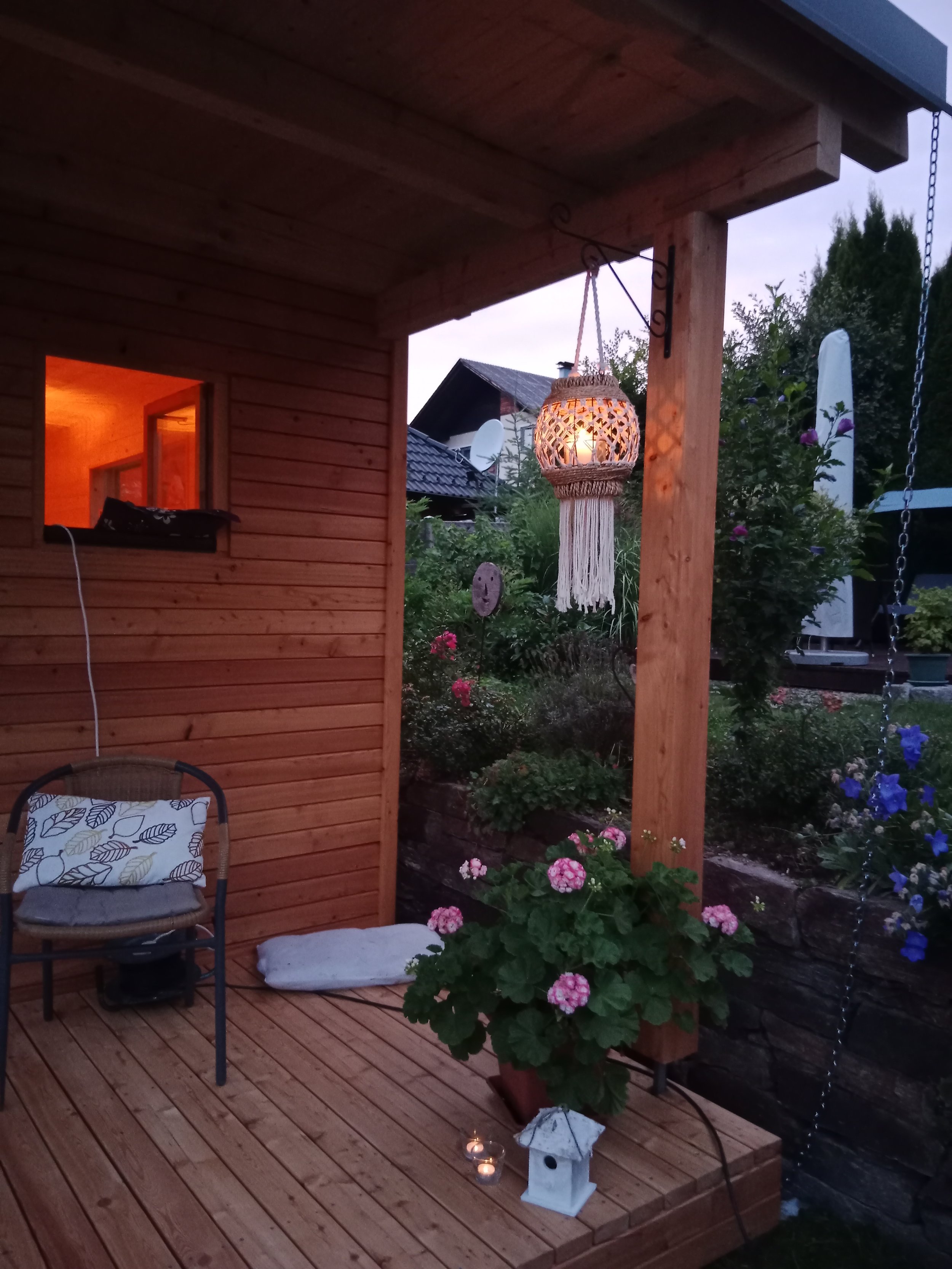 Gemütliche Laube mit Holzwänden und -boden, Beleuchtung, Garten mit bunten Blumen und Dekorationen, including einer Hängeleuchte mit Makramee, einer kleinen Laterne und einer Gartenlampe.