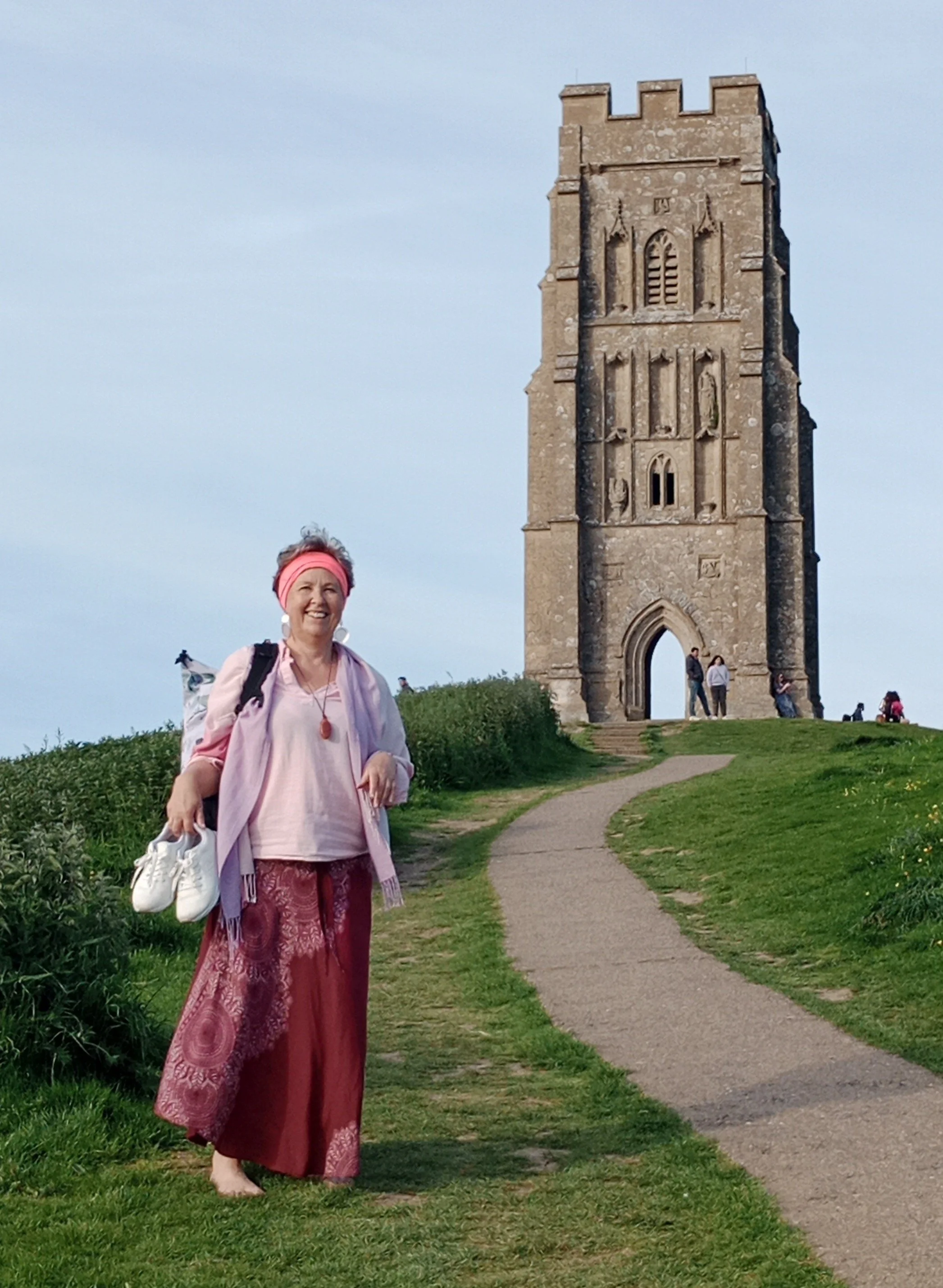 Eine Frau in pinker Kleidung steht auf einem grasbewachsenen Weg vor einem mittelalterlichen Turm, der sich auf einem Hügel befindet, umgeben von mehreren Menschen.