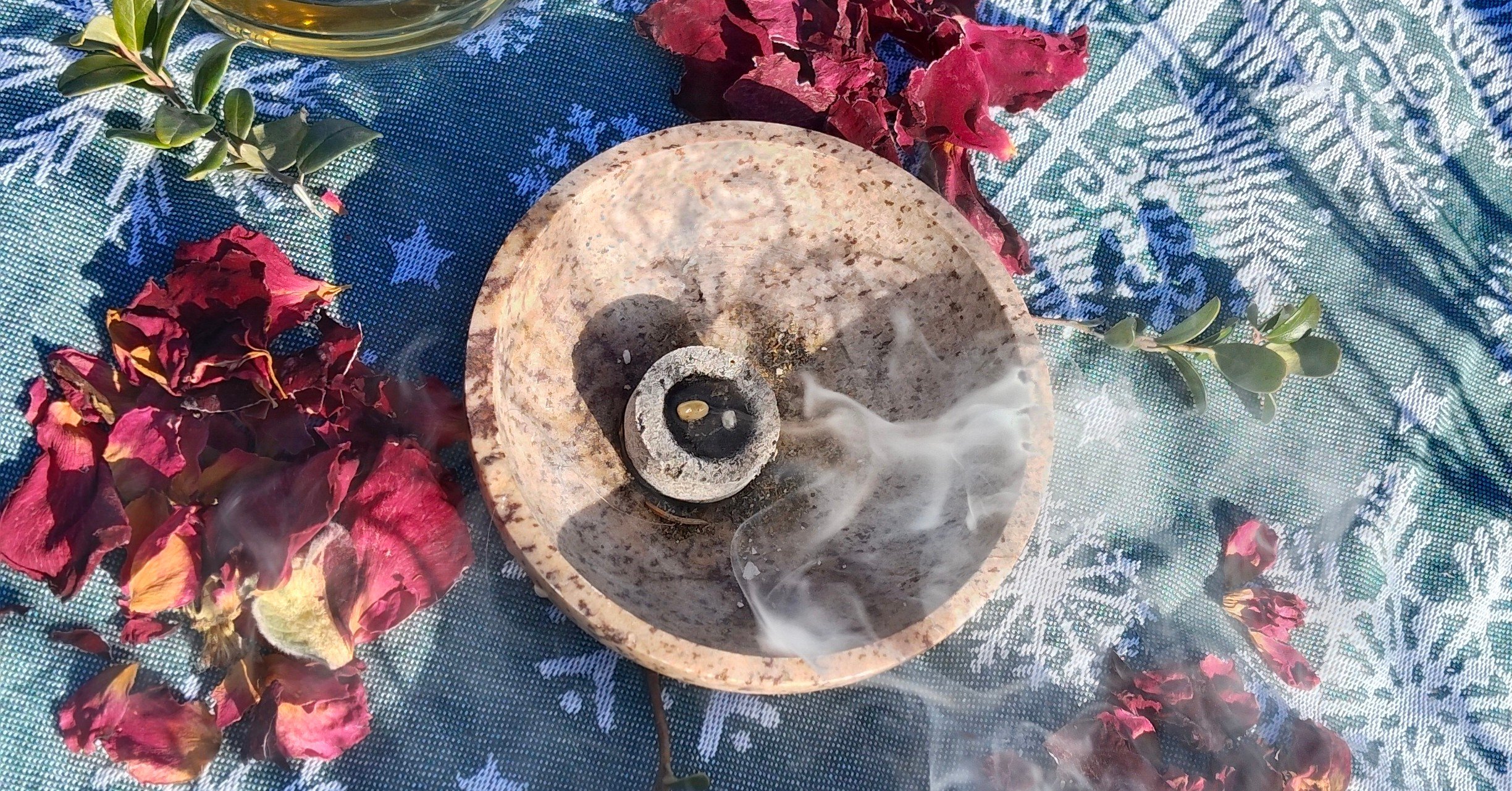 Rauchenden Räucherstäbchen auf einem Steinuntersetzer, umgeben von getrockneten Rosenblättern und grünen Zweigen, auf einer blauen Tischdecke mit weißen Blättermotiv.
