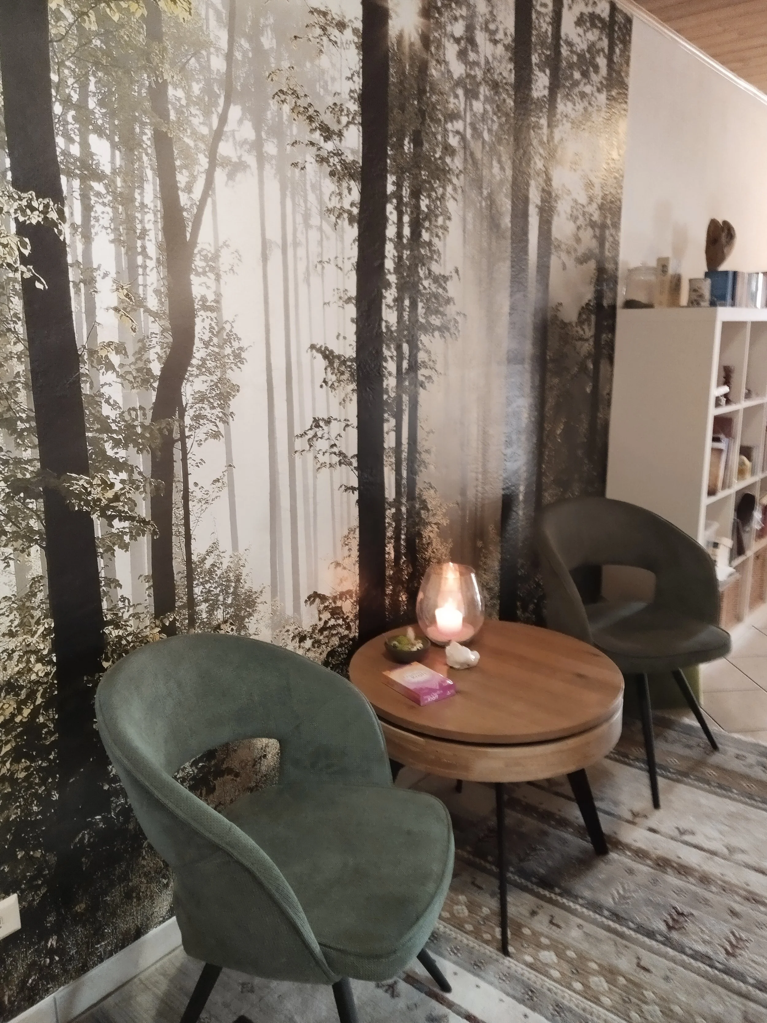 Ein gemütliches Wohnzimmer mit einer Wald-Wandtapete, einem kleinen runden Holztisch, zwei schwarzen Sesseln, einer Kerze auf dem Tisch, und einem Bücherregal im Hintergrund.