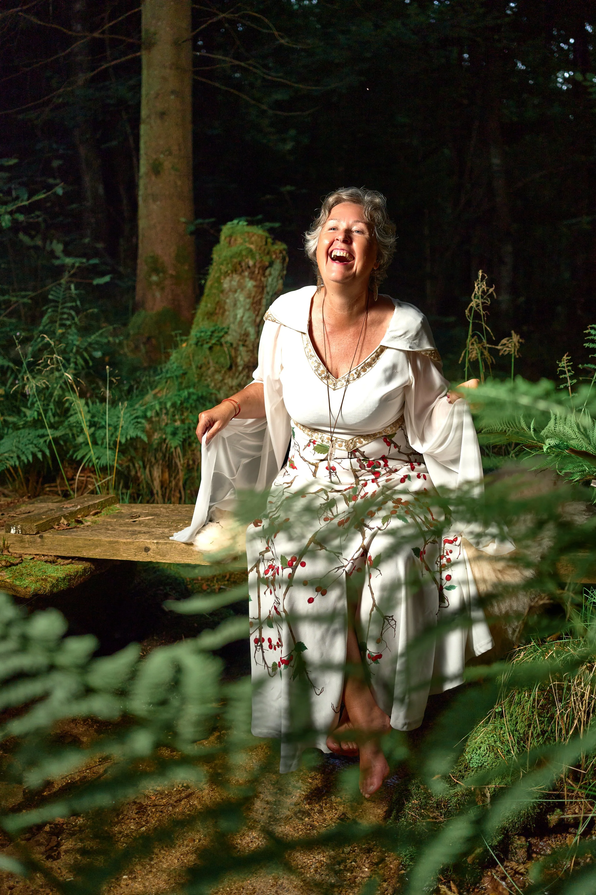Eine lachende alte Frau in einem weißen Kleid mit roten Beerenmustern sitzt barfuß auf einem Baumstamm in einem dunklen, grünen Wald.
