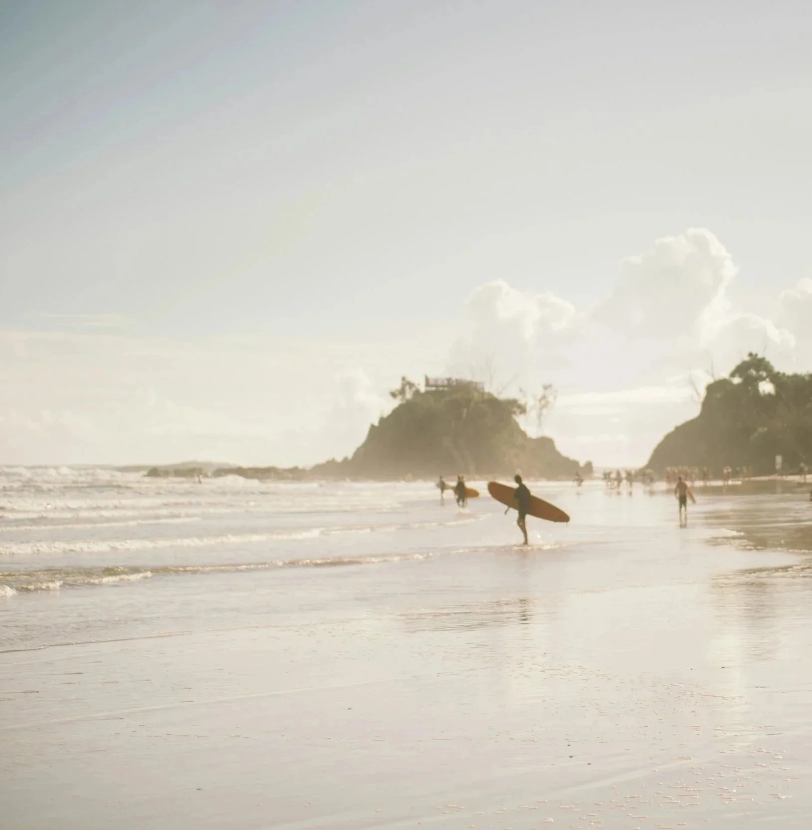 Beach scene with surfers and people walking along the shoreline, large rocky formations in the background, and a partly cloudy sky.