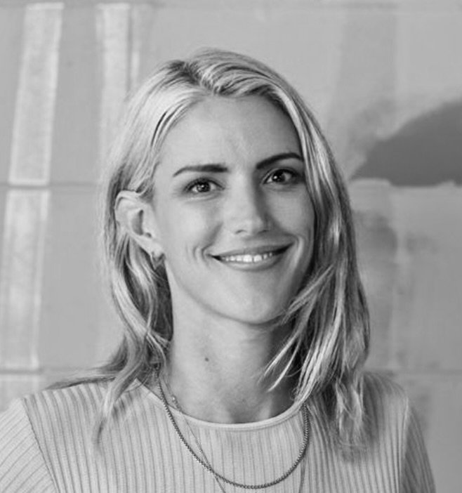 A smiling woman with shoulder-length blonde hair, wearing a light-colored top and a necklace, standing in front of a brick wall.