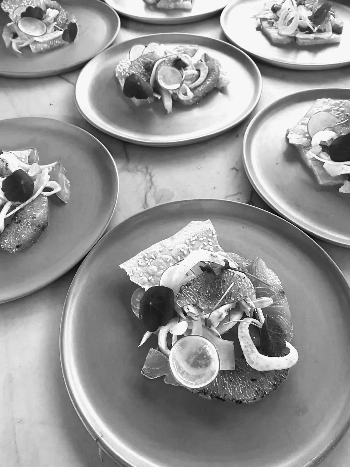 Several plates of appetizers or small dishes, each with a piece of toast topped with various vegetables, citrus slices, and garnishes, arranged on a marble table.