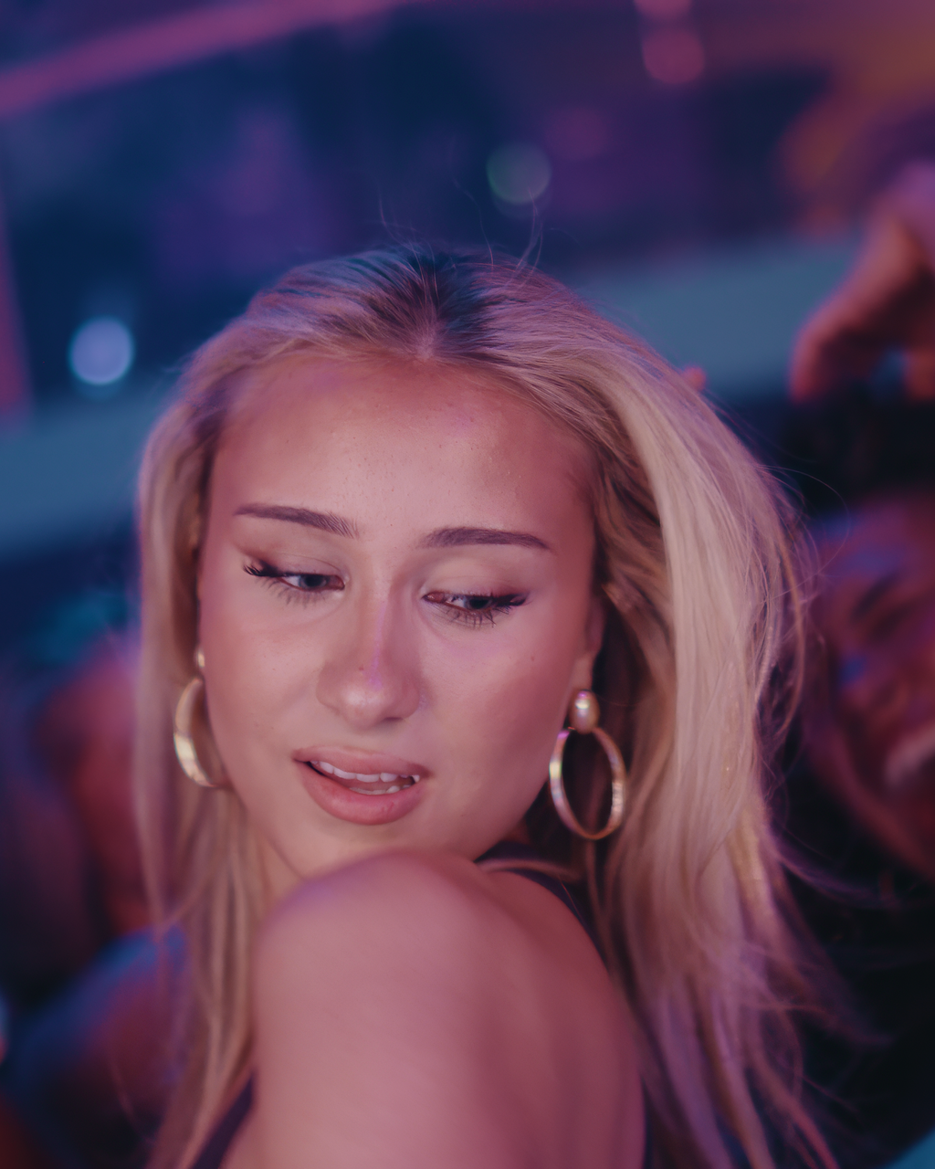 Close-up of a young woman with blonde hair, wearing hoop earrings, looking down with a playful expression in a colorful, blurry background.