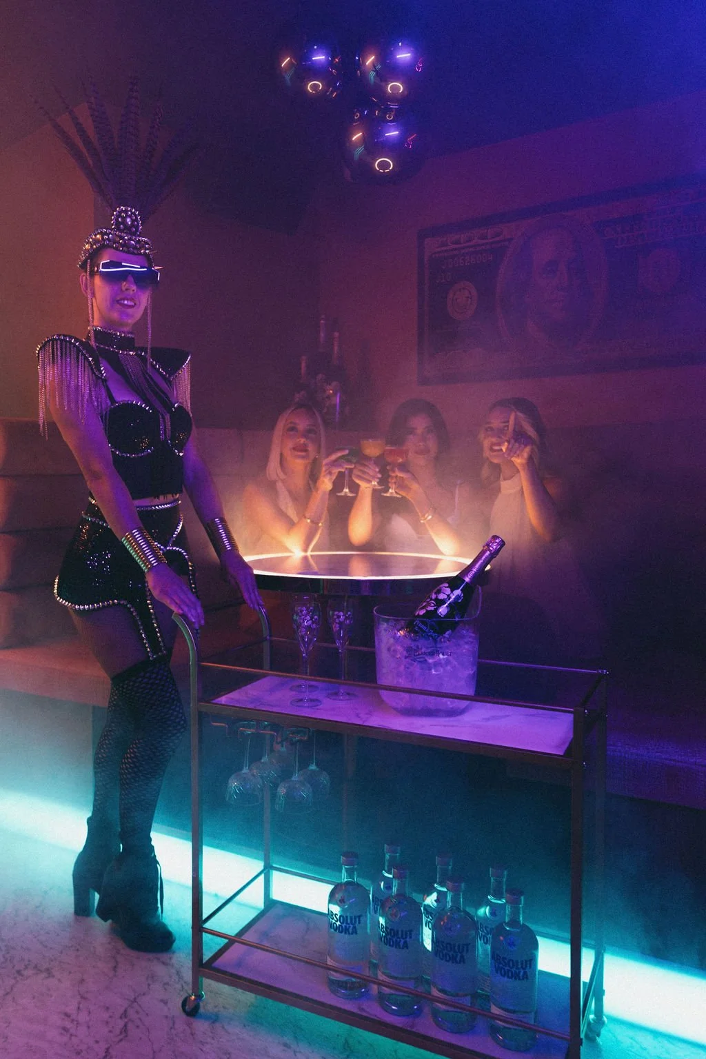 A themed party scene with a performer in futuristic attire and three women in a nightclub, holding cocktails and celebrating with a large bottle of Absolut Vodka in an ice bucket, amid neon lights and a large dollar bill poster.