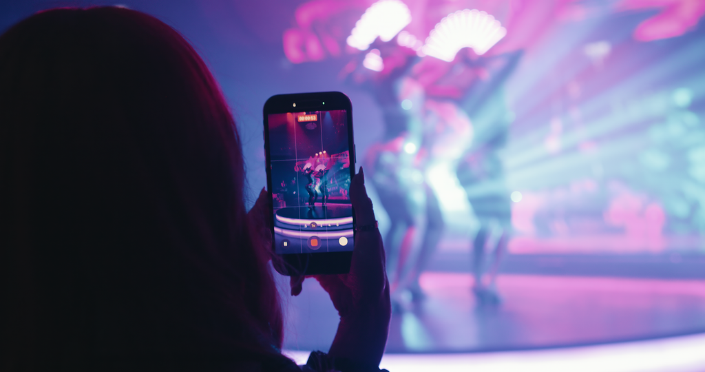 Person taking a photo or recording video of dancers on stage with colorful lighting.