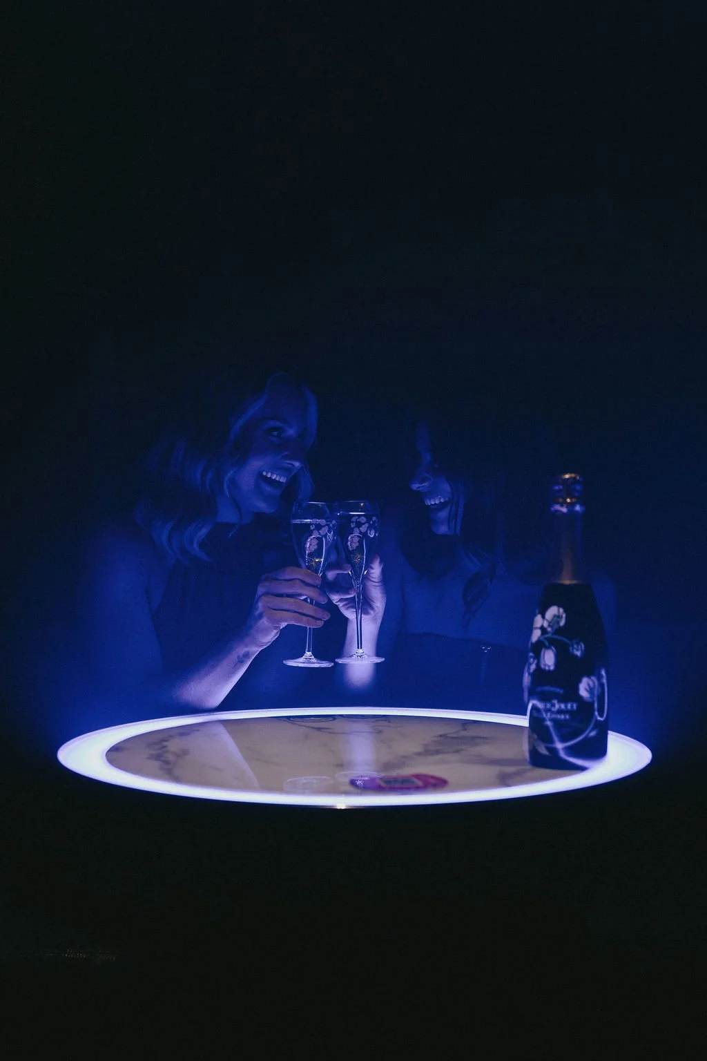 Two women smiling and_toasting with champagne glasses in a dark room with a glowing round table and a bottle of champagne.
