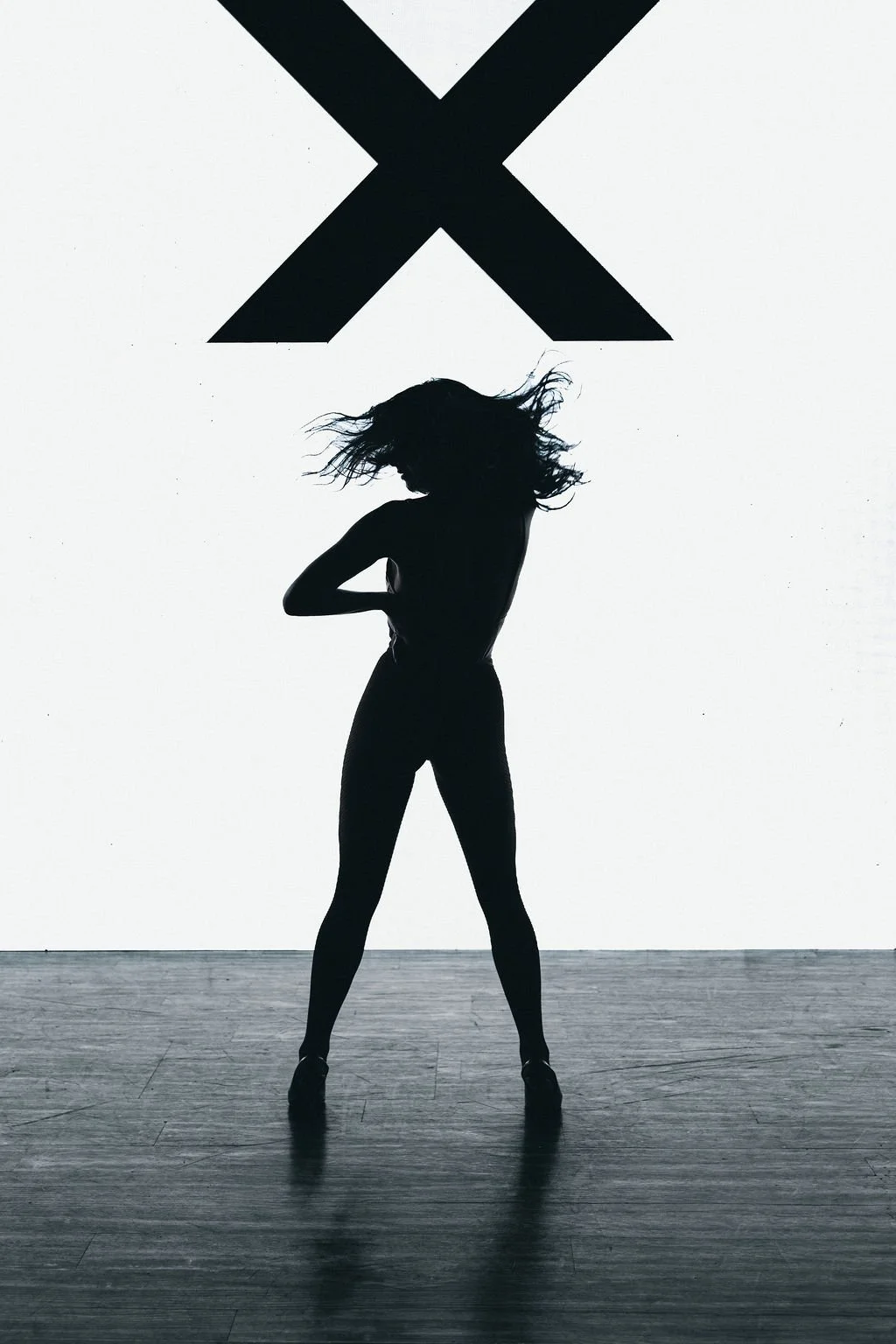 Silhouette of a woman with long flowing hair standing on a stage with a large 'X' symbol in the background.