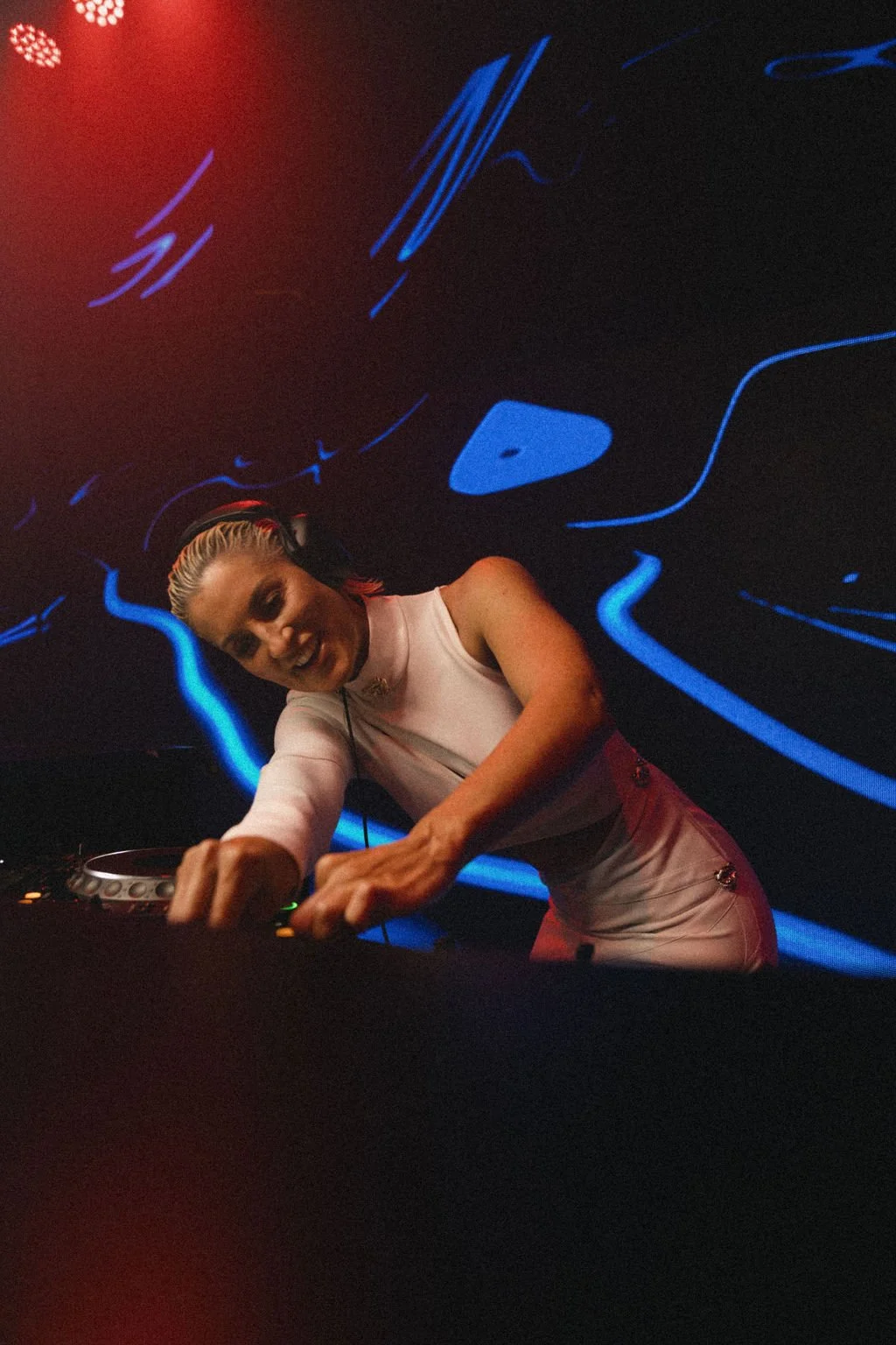 A female DJ wearing headphones, mixing music at a club with vibrant blue neon lights and digital designs on the wall behind her.