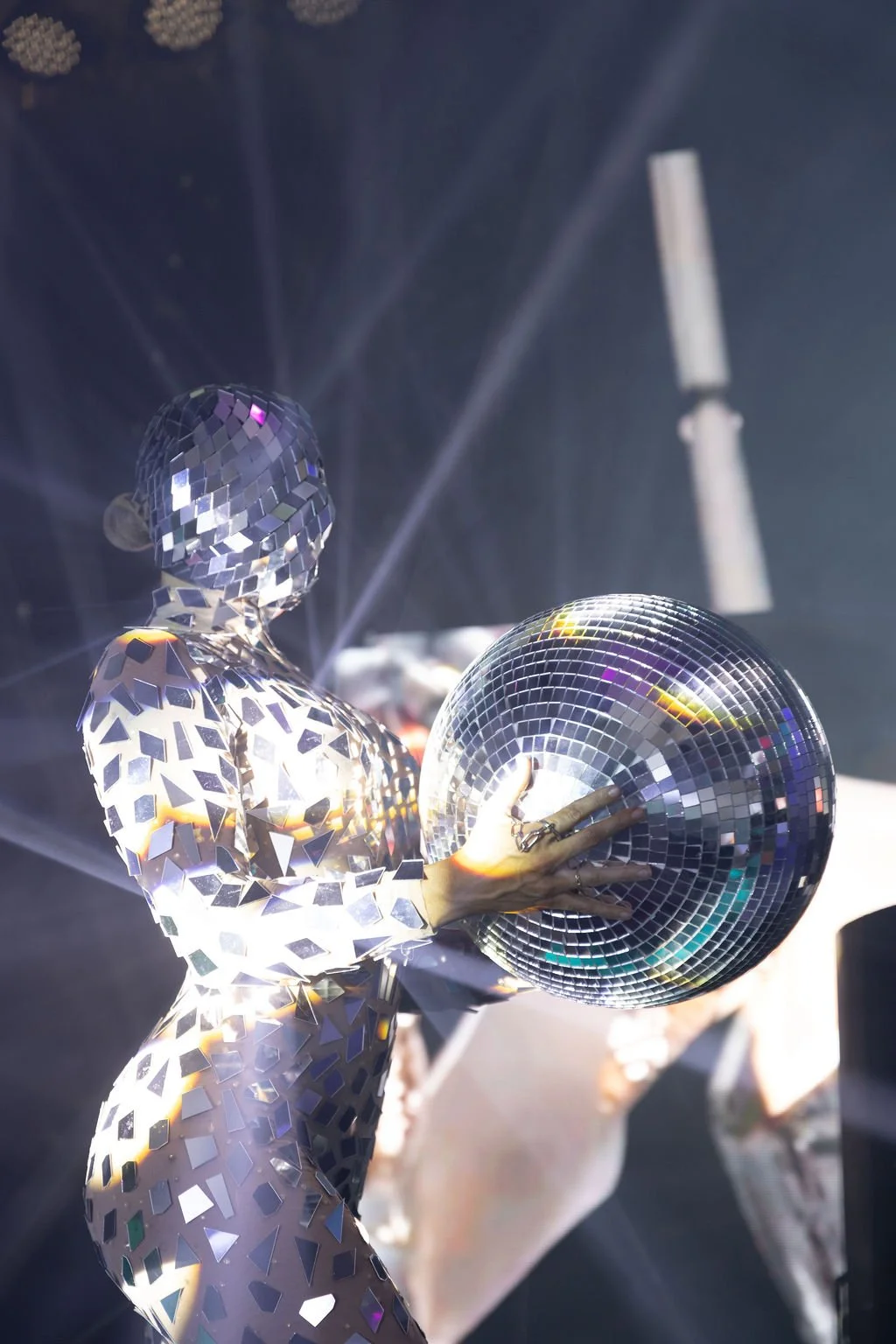 A person in a mirrored, mosaic-style suit holding a disco ball in a dark setting with colorful reflections.