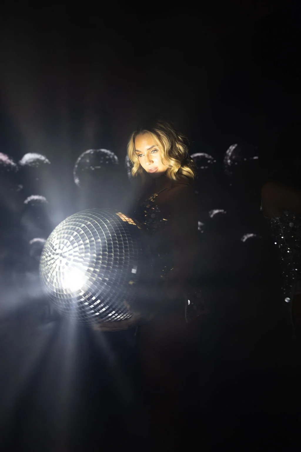 A woman with blonde wavy hair and makeup standing in a dark room with disco balls reflecting light, holding a large disco ball.
