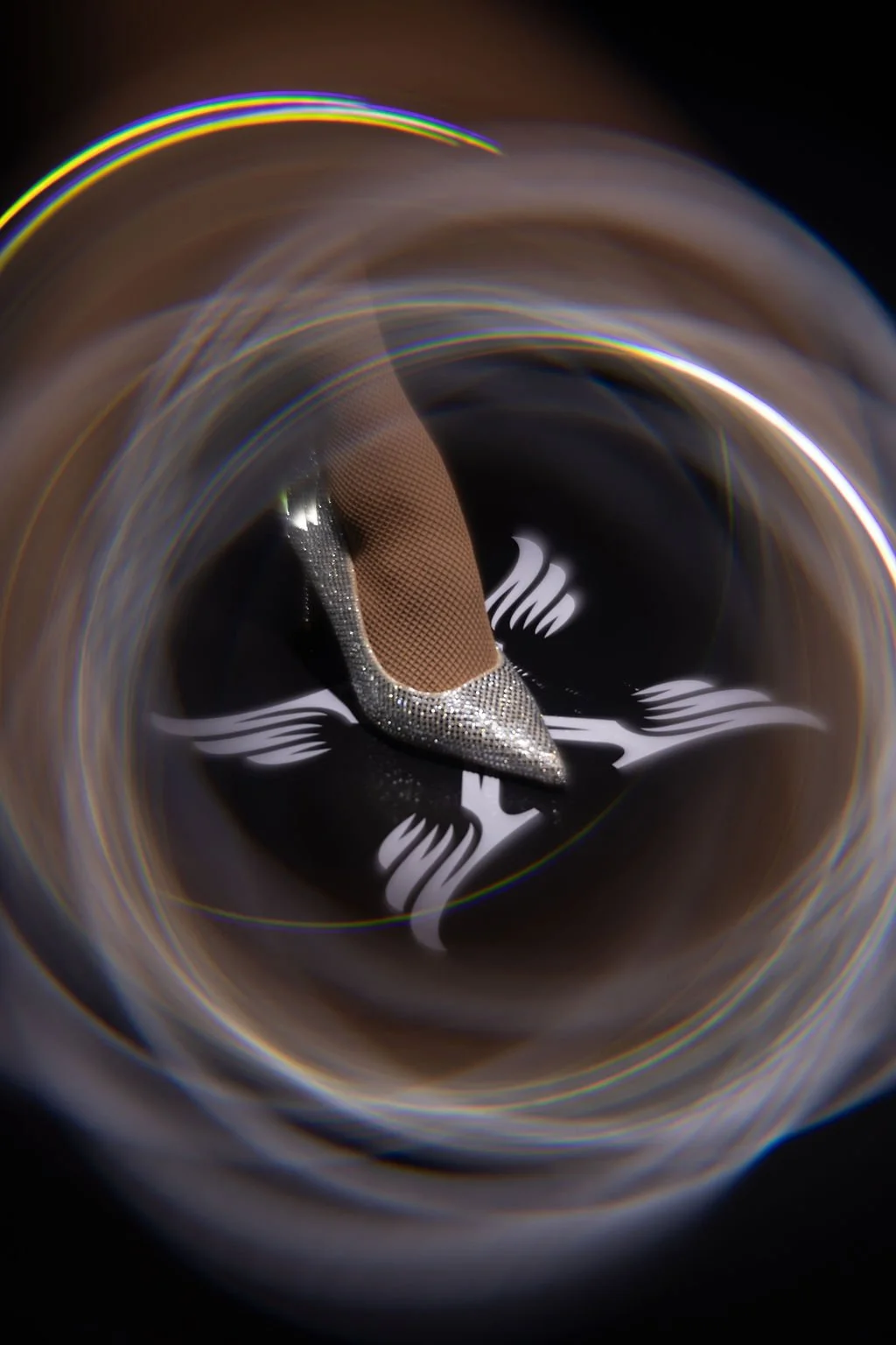 Close-up of a high-heeled shoe with rhinestones, placed on a reflective black surface with decorative white design, viewed through a circular, rainbow-like lens.