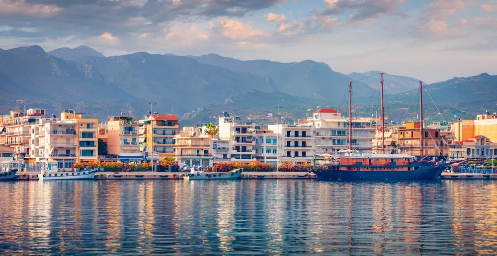 A waterfront view of Kalamata marina, in Messenia, with boats and a large sailboat, in front of a row of colorful apartment buildings with mountain scenery in the background.