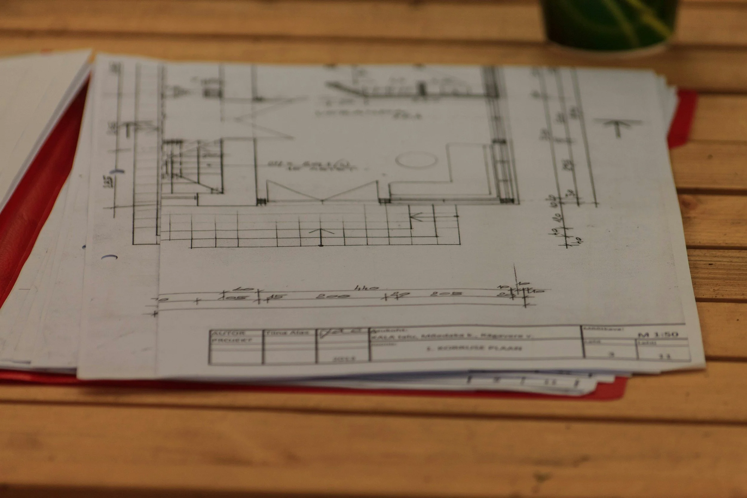 A printed architectural or engineering blueprint of a building layout placed on a wooden surface, with a partially visible plant in a green container in the background.