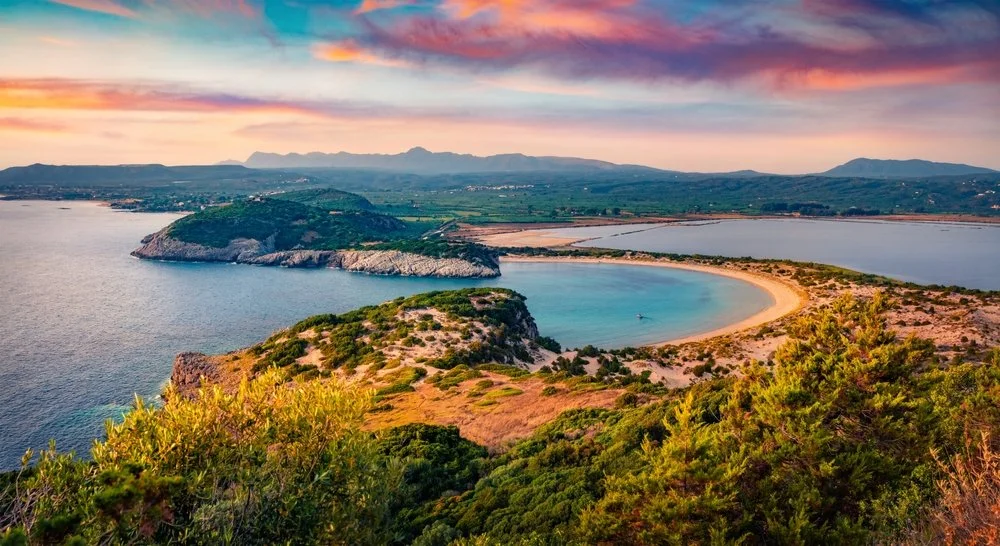 A panoramic view of Voidokilia beach, a sandy beach and horseshoe shaped cove, near Gialova, Messenia, surrounded by lush green hills and distant mountains under a colorful sunset sky.