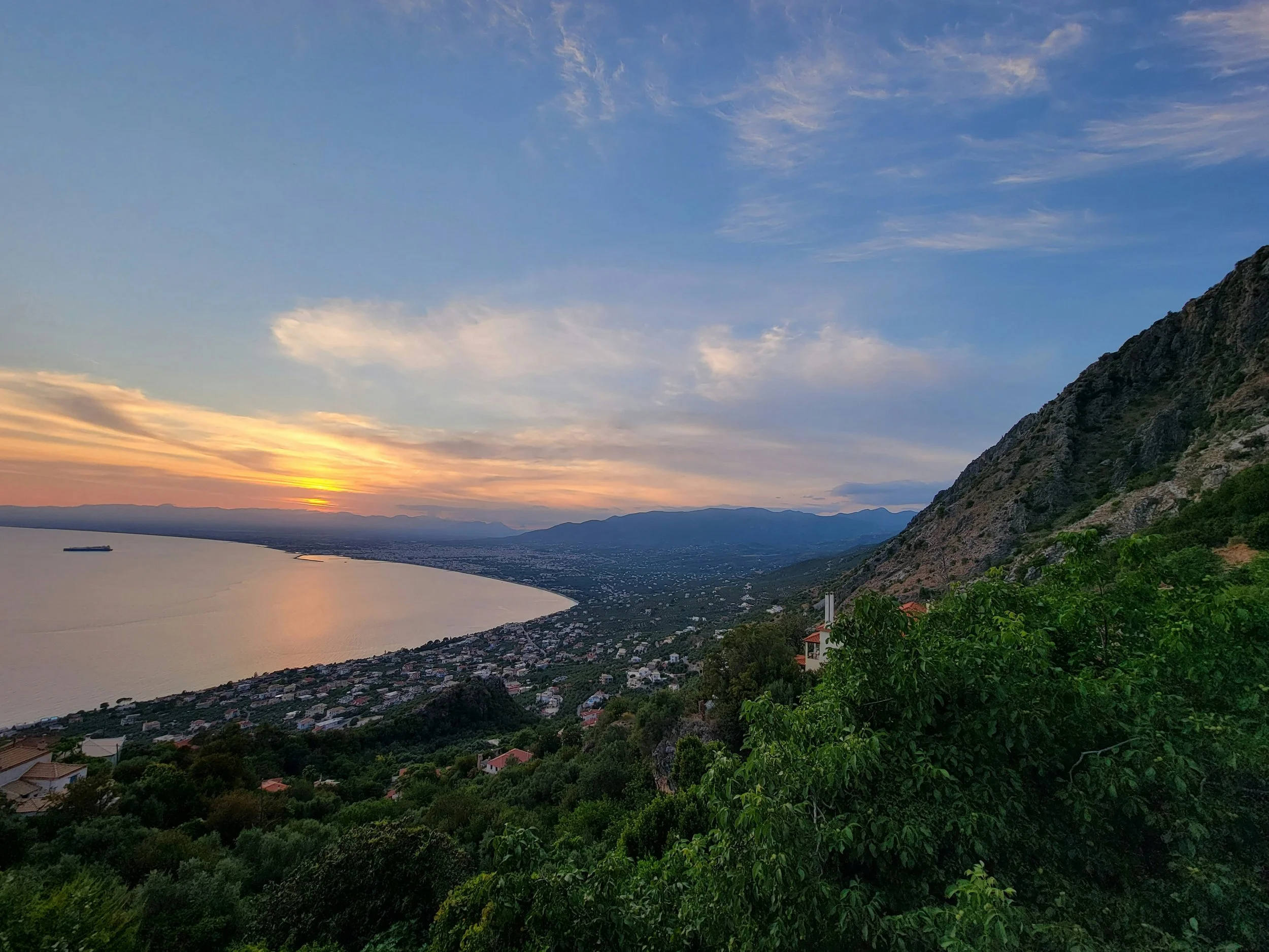 A scenic view of the Messenian Gulf , looking from Verga towards Kalamata, with mountains in the distance, captured at sunset with colorful sky.
