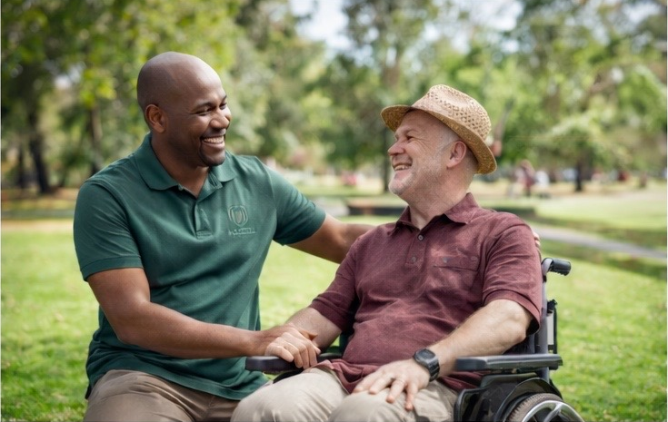 A man in a green polo shirt is smiling and interacting with a man in a wheelchair who is wearing a maroon shirt and a straw hat, with a park and trees in the background.