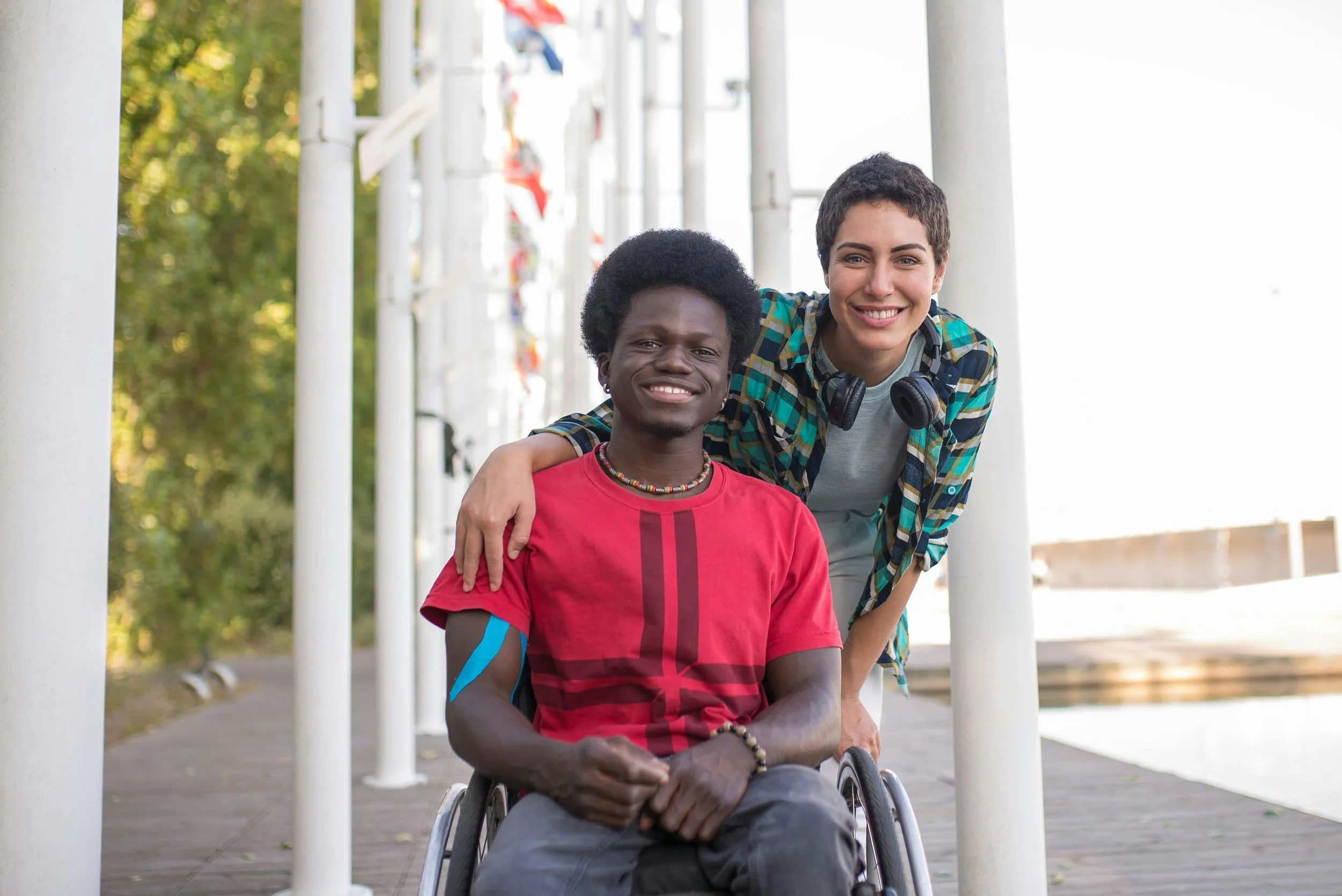 A person in a wheelchair and a woman with short dark hair are smiling and posing together outdoors, with the woman resting her arm on the person's shoulder.