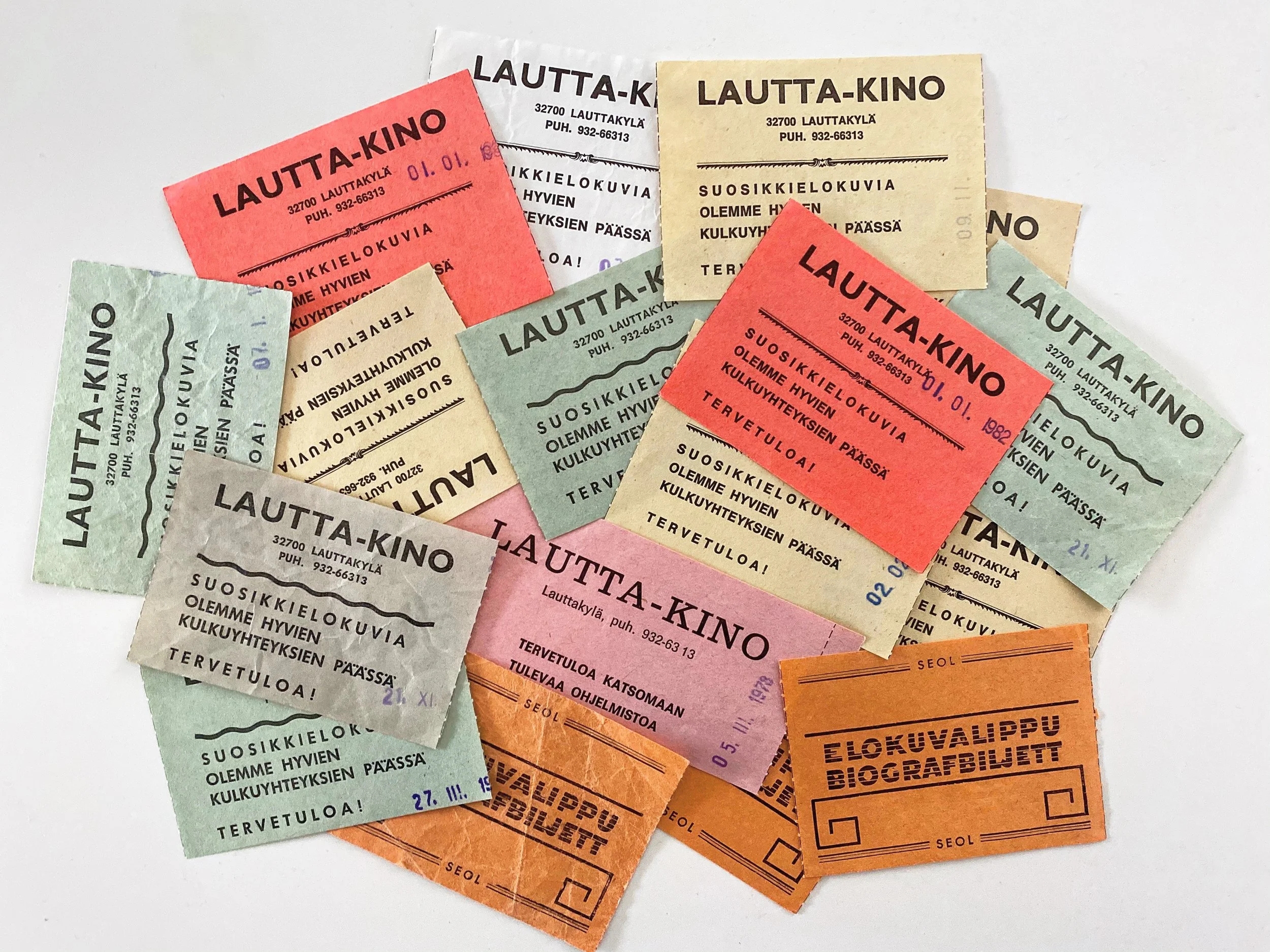 Useita epäyhtenäisiä lippuja ja lappuja, joista osa on punaisia, vihreitä, keltaisia, beigejä, pinkkejä ja oransseja, mainostaen Lauttakylän elokuvateatteria.