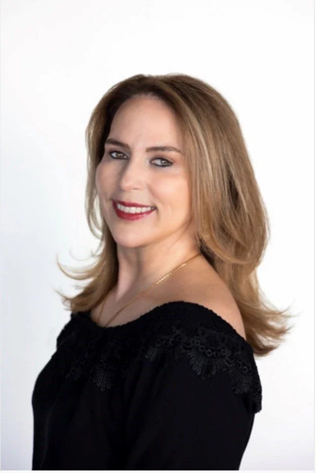 A woman with shoulder-length blonde hair, smiling, wearing a black off-the-shoulder top and a gold necklace, against a white background.
