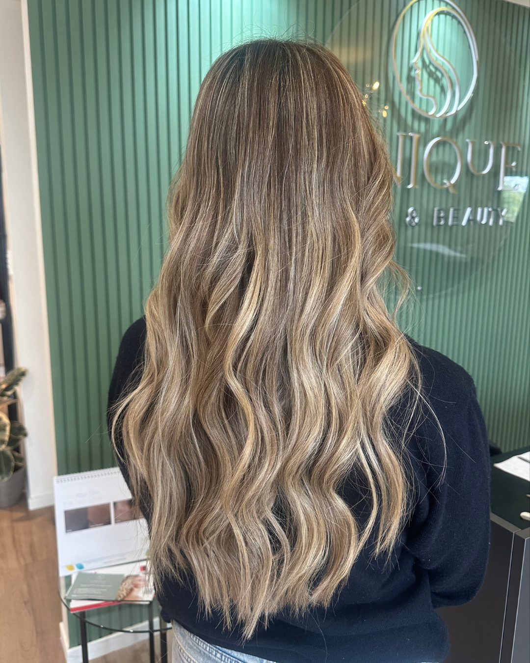 Back view of a woman with long, wavy blonde hair inside a salon with green walls marked 'L'Q' and 'BEAUTY' in the background.