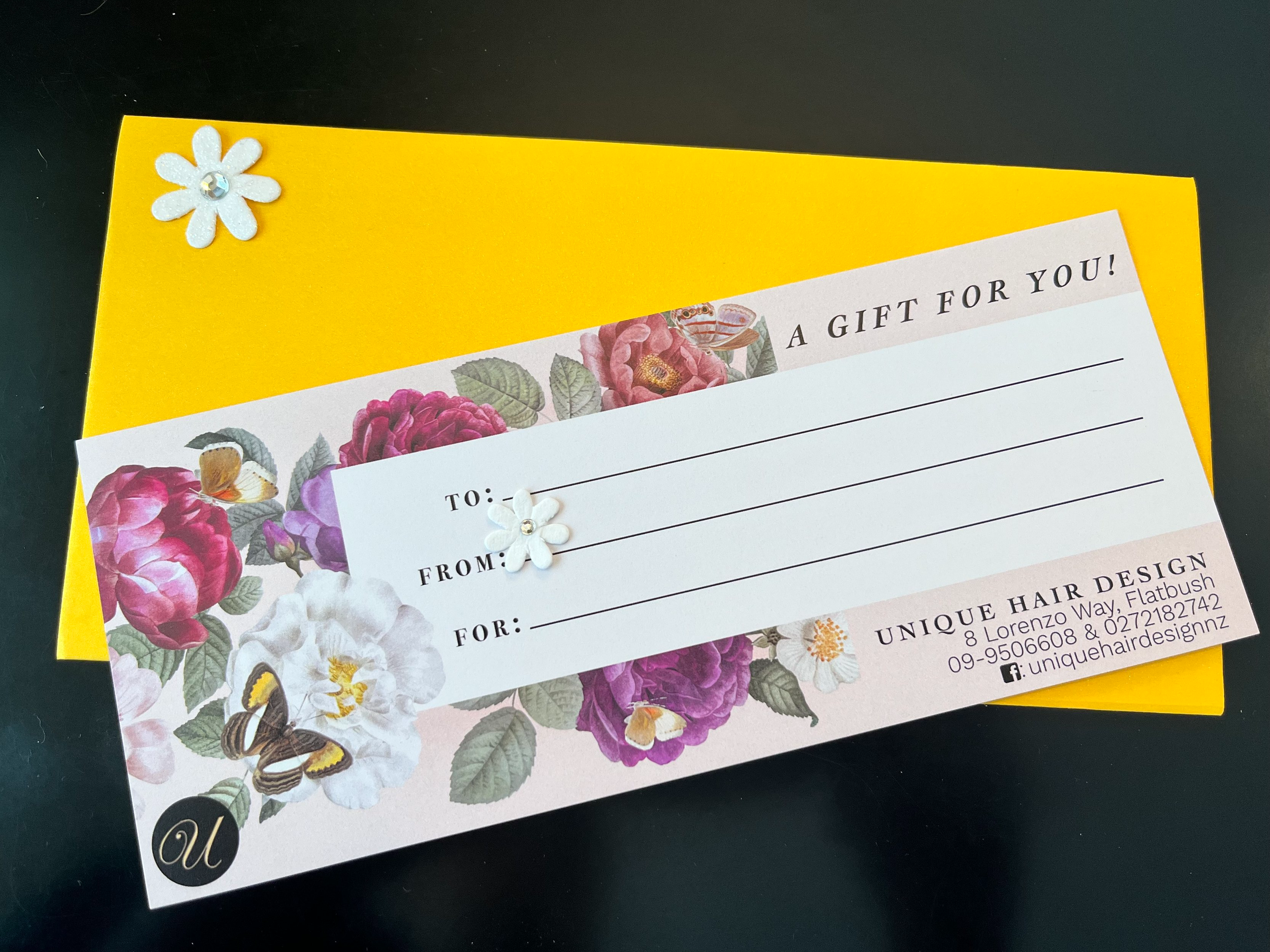 Part of a floral-themed gift voucher with a yellow background, featuring colorful flowers and butterflies, and decorative white foam flowers with rhinestones. The voucher includes spaces for 'To,' 'From,' and 'For' information.