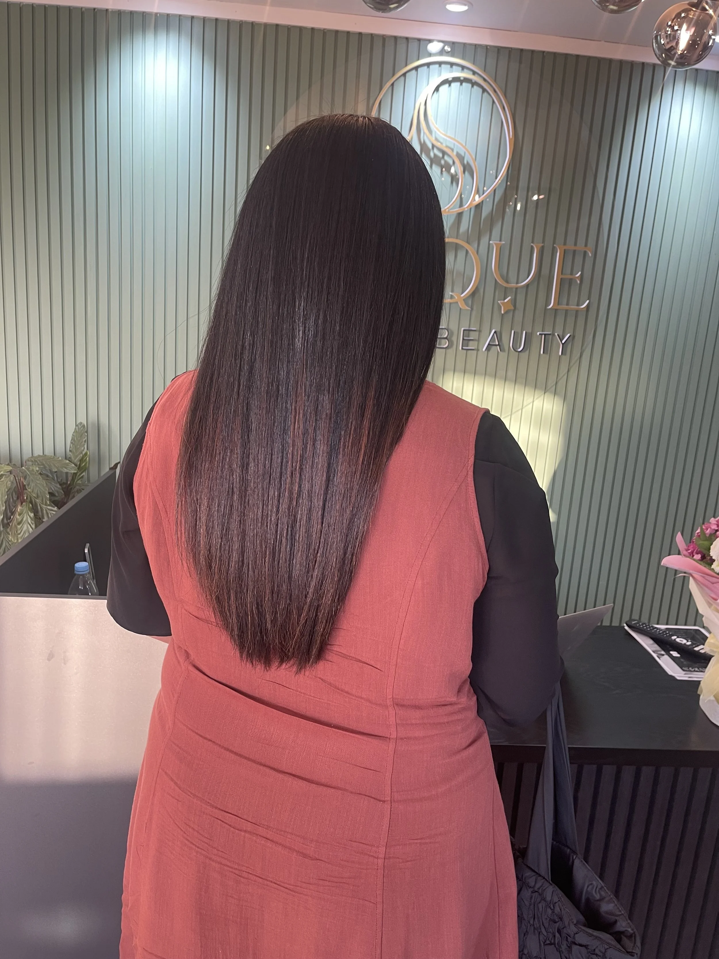 Woman with long, straight, dark brown hair wearing a red and black outfit standing in front of a sign that reads 'DUE BEAUTY' at a beauty salon or spa.