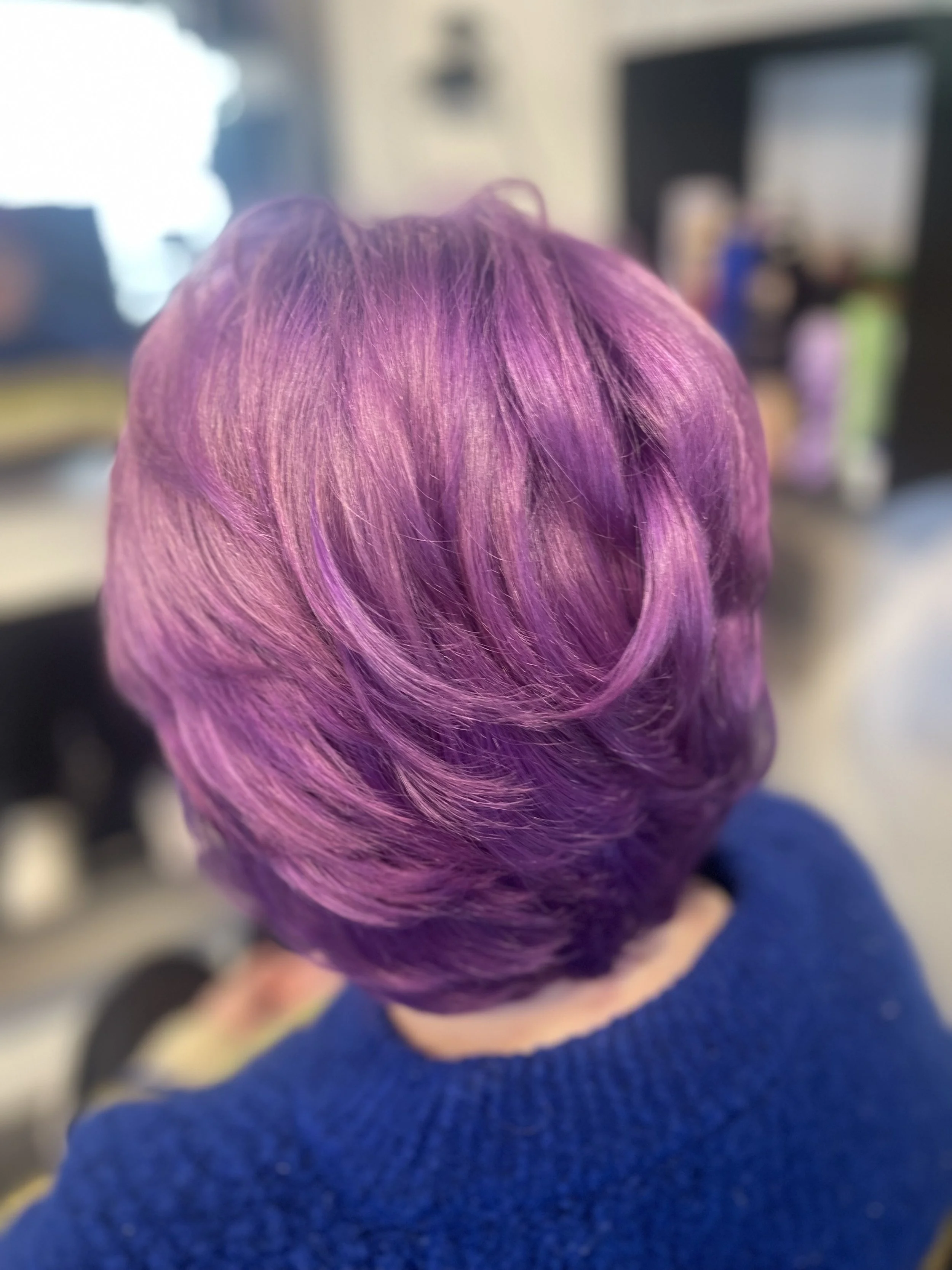 Close-up of a person's head with short hair dyed in shades of pink and purple, wearing a blue sweater.