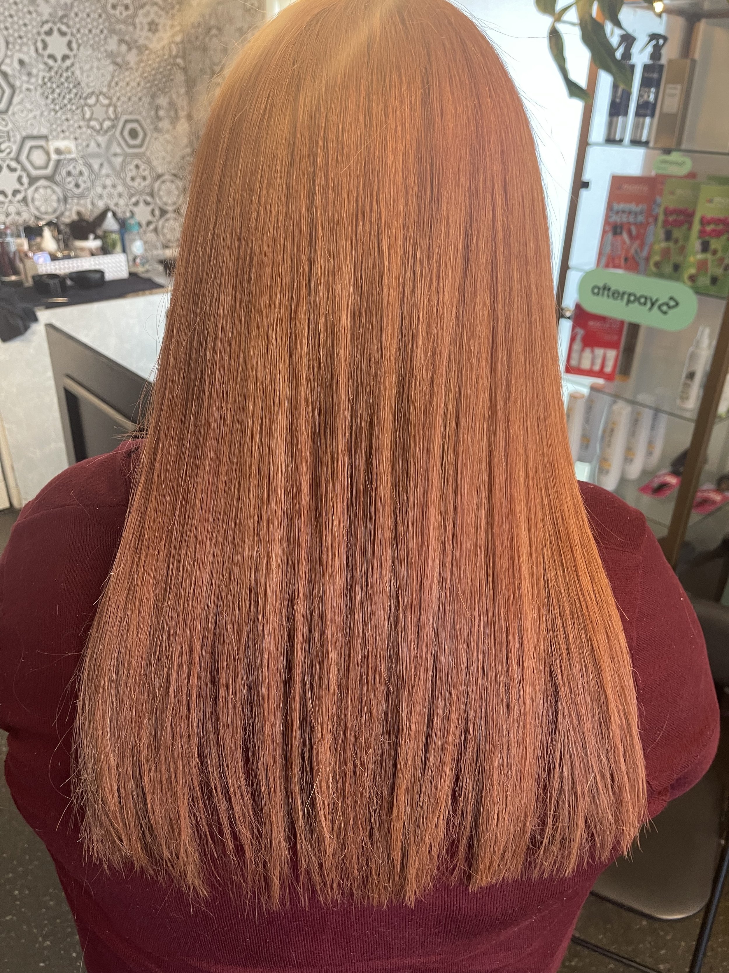 Close-up of a person with straight, shiny, orange-red hair, wearing a dark maroon top, in a salon or store setting.