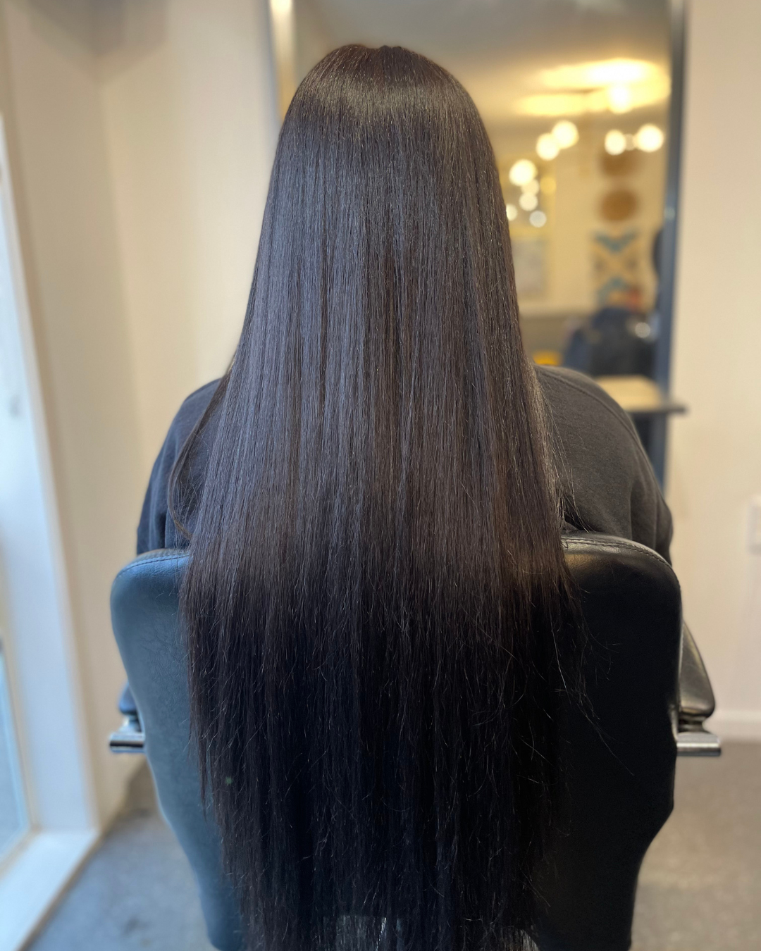 Back view of a woman with long, straight, shiny black hair sitting in a salon chair.