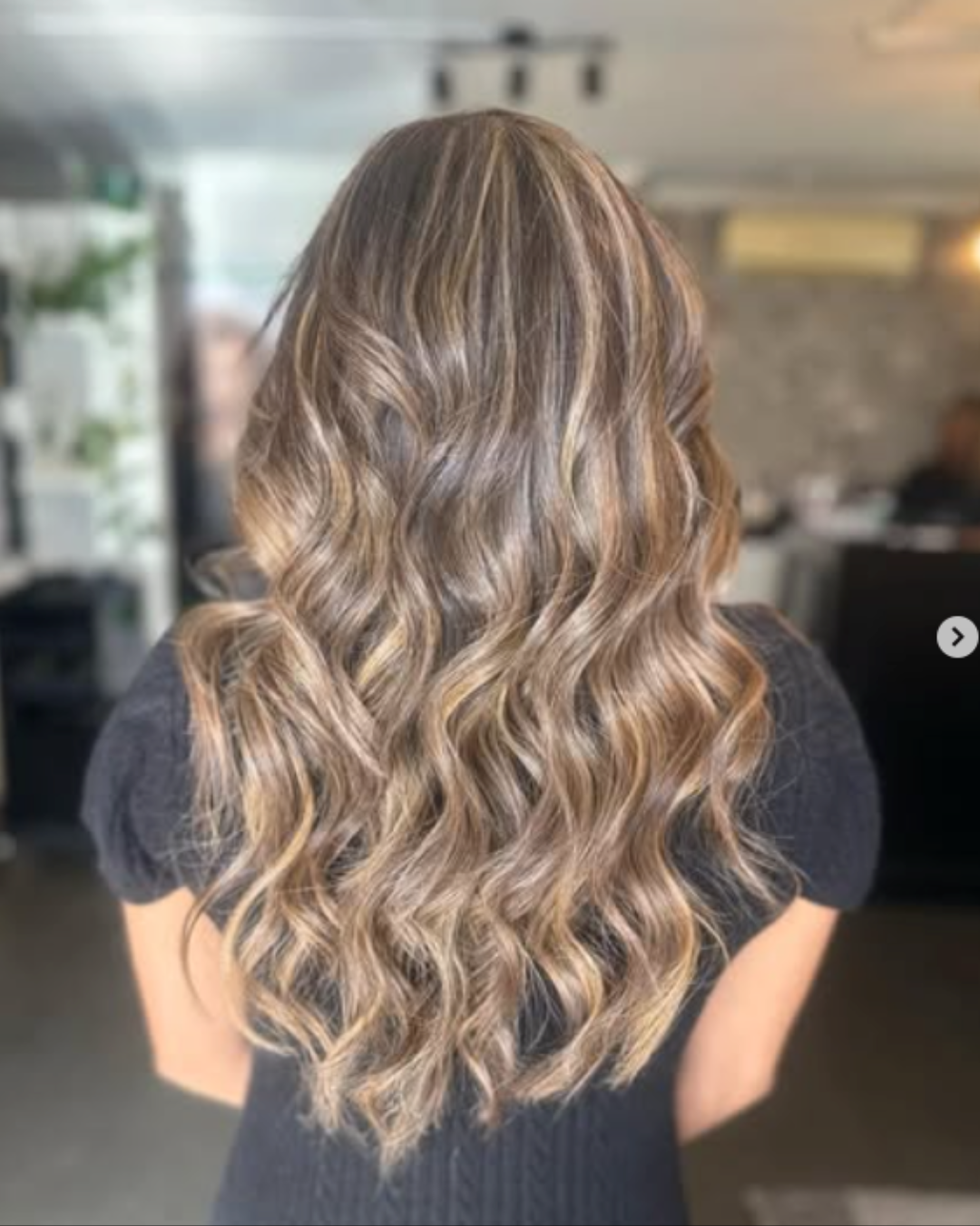 Back view of a woman with long, wavy, blonde hair with highlights, indoors in a salon or similar setting.