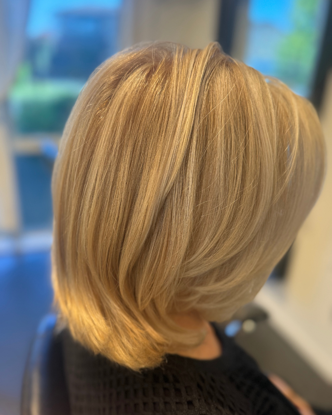 A woman with shoulder-length blonde hair styled with soft layers, seen from behind, sitting indoors near a window with natural light.