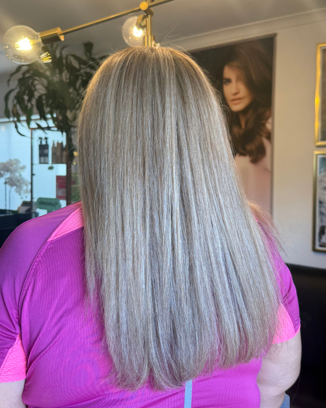 Back of a woman with long, straight blonde hair wearing a pink shirt in a salon with framed pictures on the wall and a large portrait of a woman with brown hair in the background.