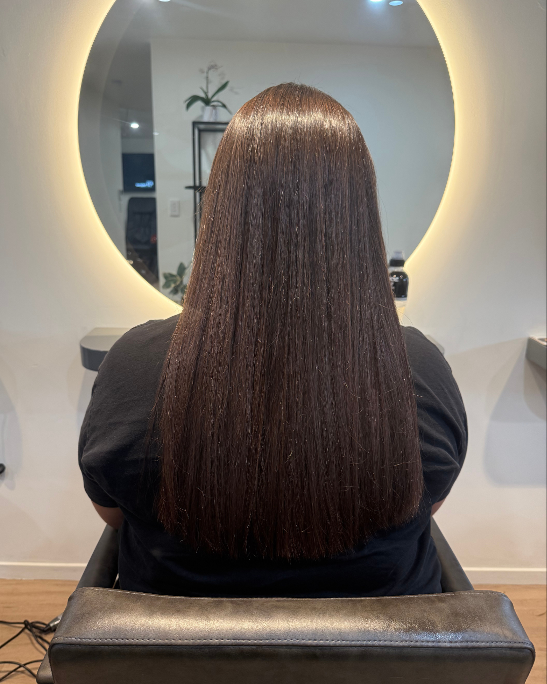Back view of a person with long, straight, dark brown hair sitting in a salon chair in front of a large oval mirror with backlit border.