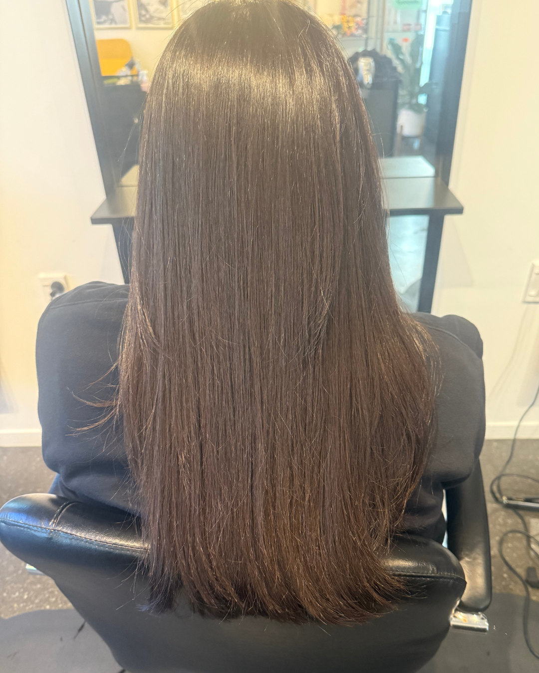 Back view of a woman with long, straight, brown hair sitting in a salon chair.