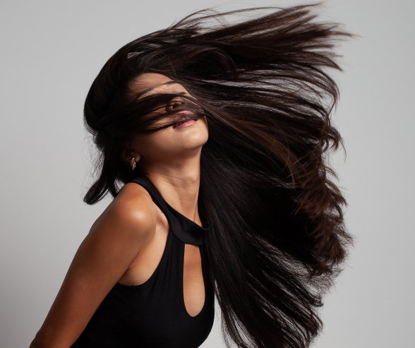 Woman with long, dark hair flipping in the air, wearing a black sleeveless top against a plain gray background.