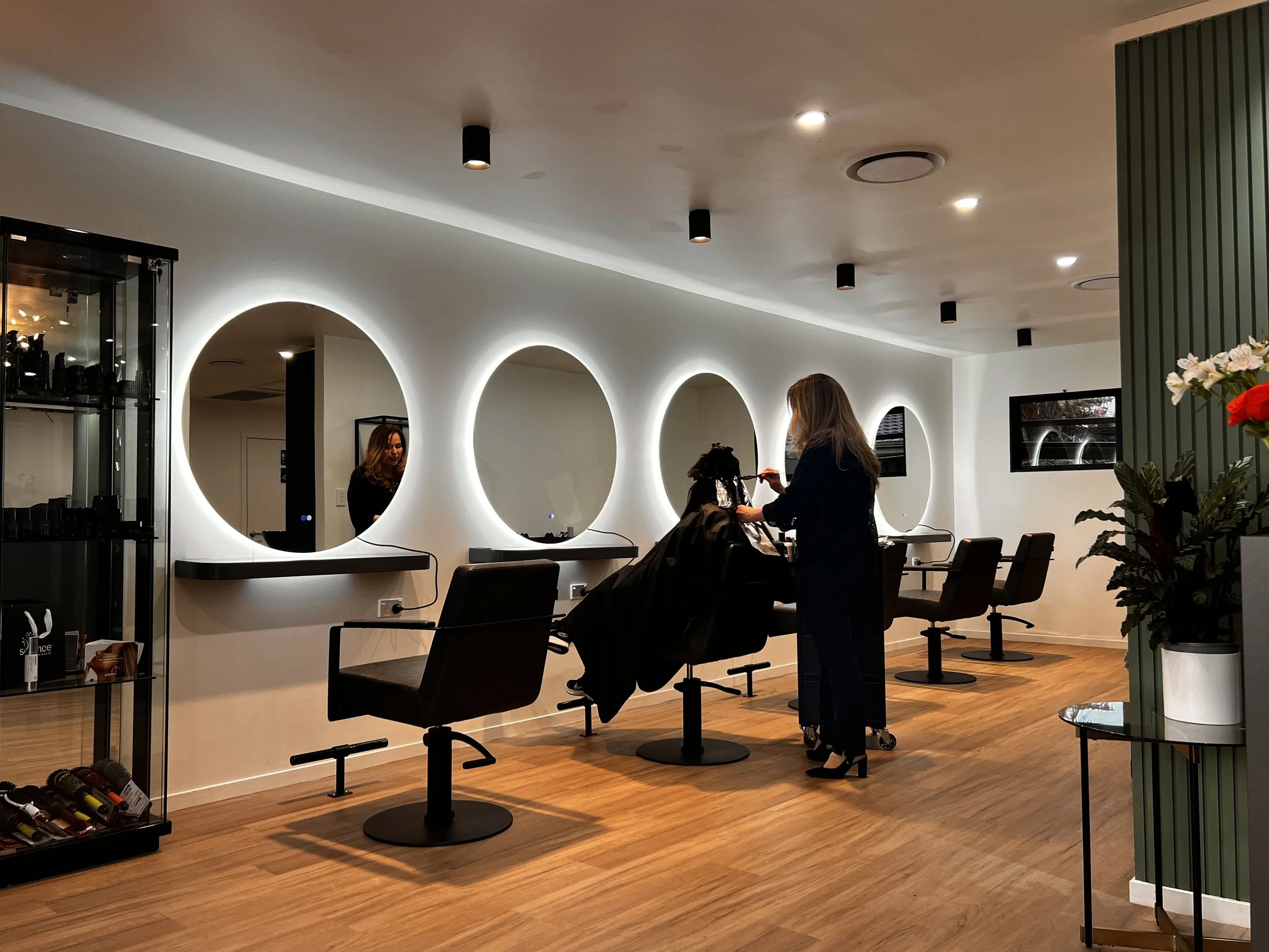 A modern hair salon interior with circular illuminated mirrors on a white wall, two women getting hair treatments, and hair styling tools on a nearby table. The space has warm wooden flooring and ceiling lights.