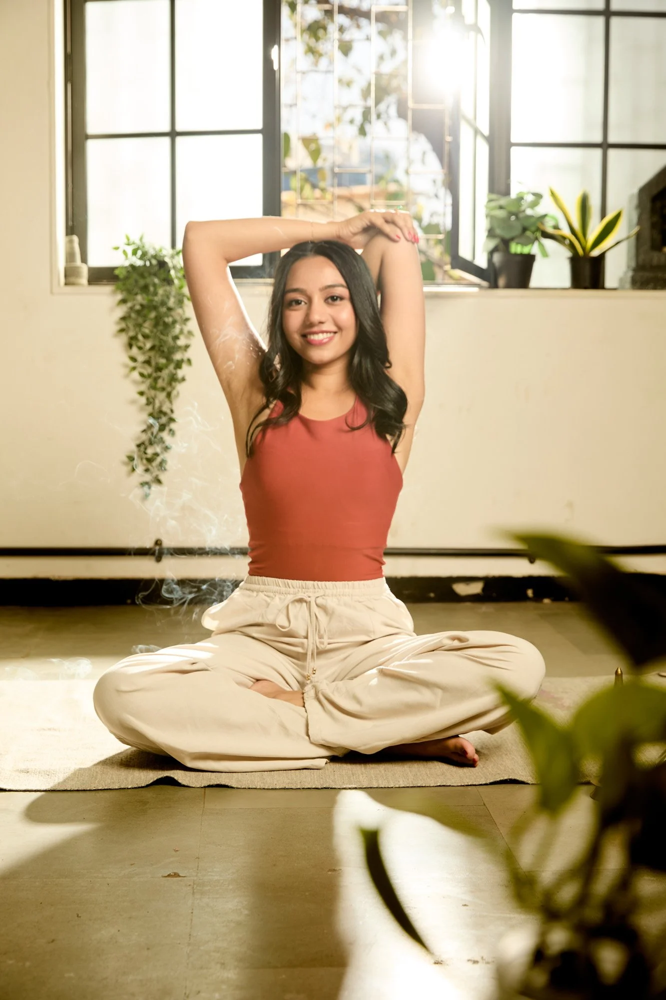 A woman sitting cross-legged on a yoga mat indoors, with her arms raised above her head, smiling, and sunlight coming through the window behind her.