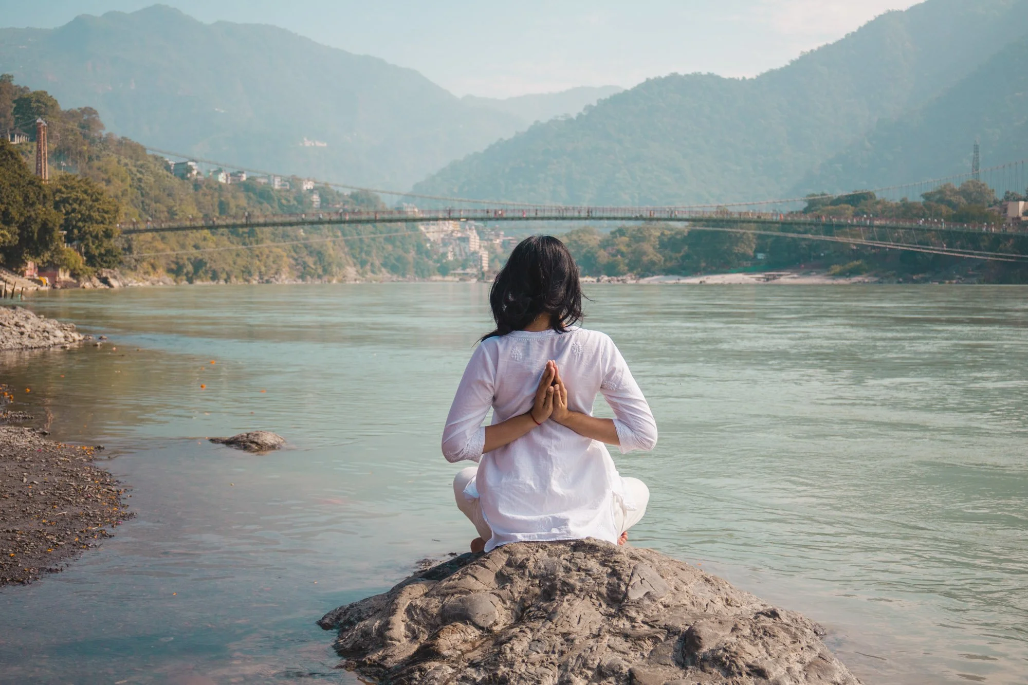 A woman practicing yoga on a rock by a river in a mountainous landscape with a suspension bridge in the background.