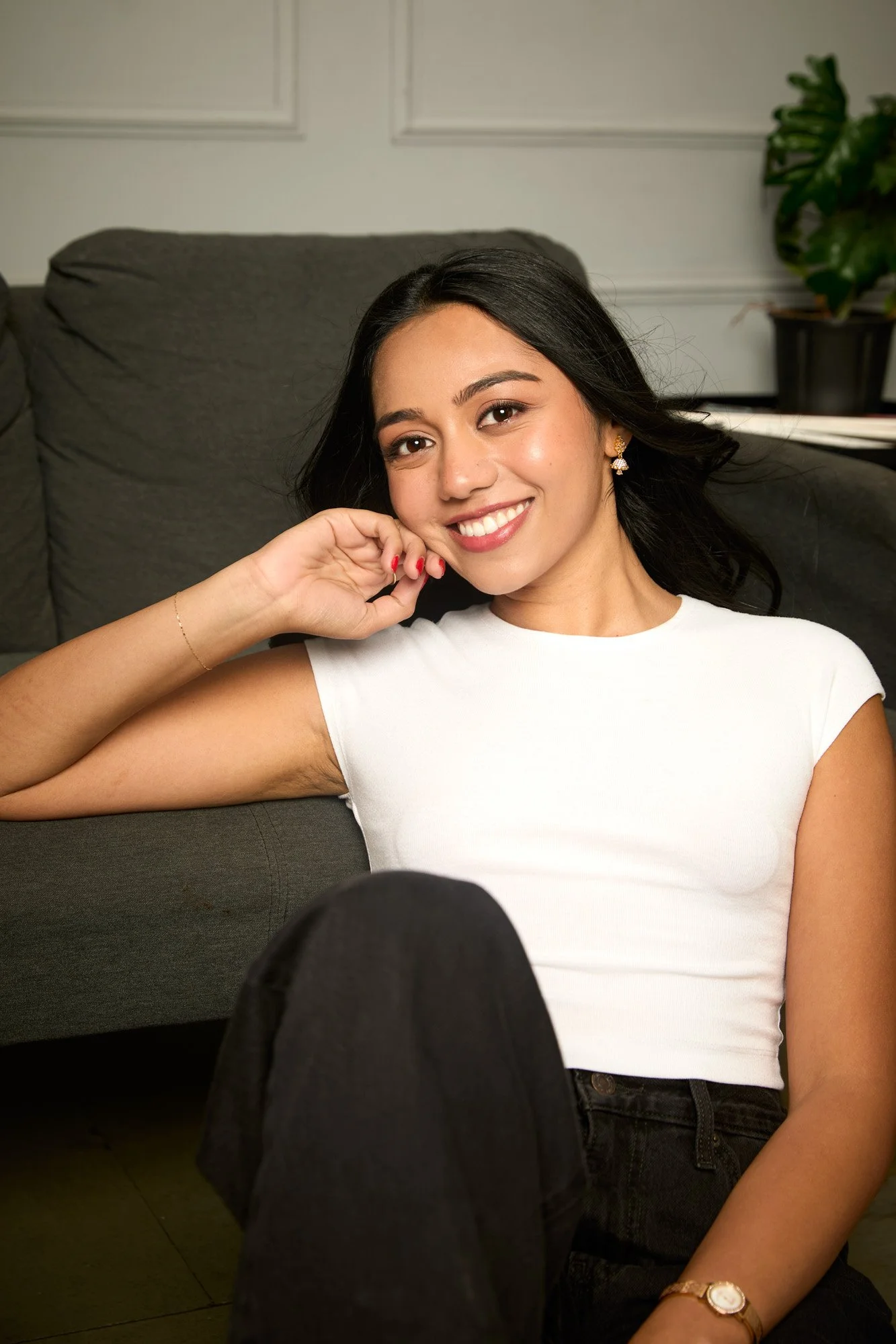 A young woman with dark hair, smiling, lying on a dark grey couch, wearing a white sleeveless top and black pants, with a plant in the background.