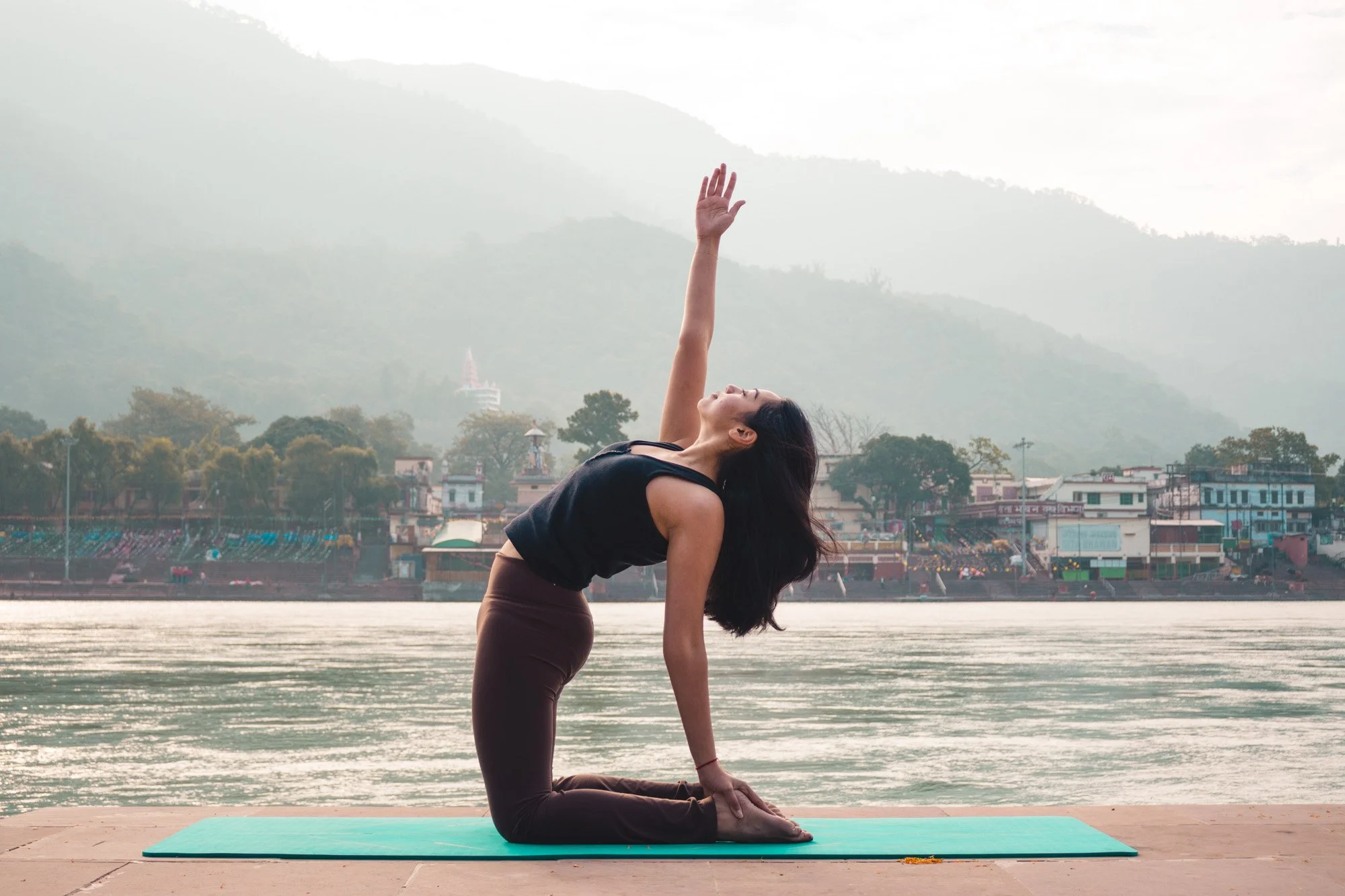 Woman practicing yoga outdoors on a mat near a river with mountains and buildings in the background.