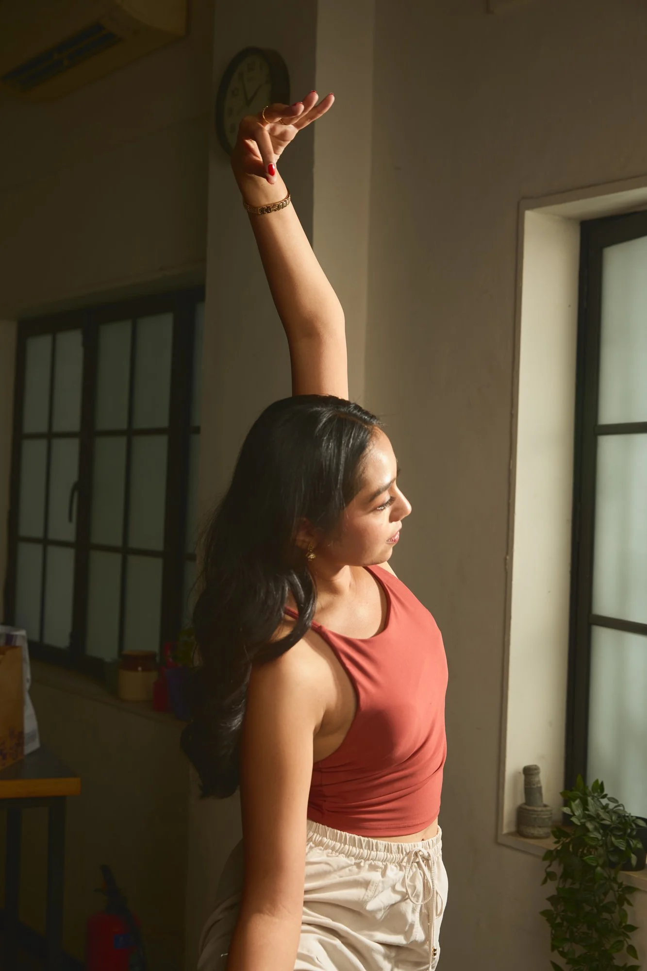 A woman with long dark hair wearing a sleeveless rust-colored top and white drawstring shorts is standing indoors near a window, reaching her arm up with her hand relaxed.