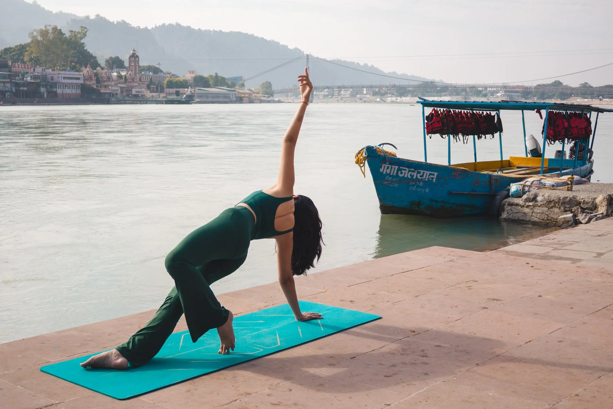 A woman practicing yoga on a blue mat on a riverside dock with a boat and cityscape in the background.